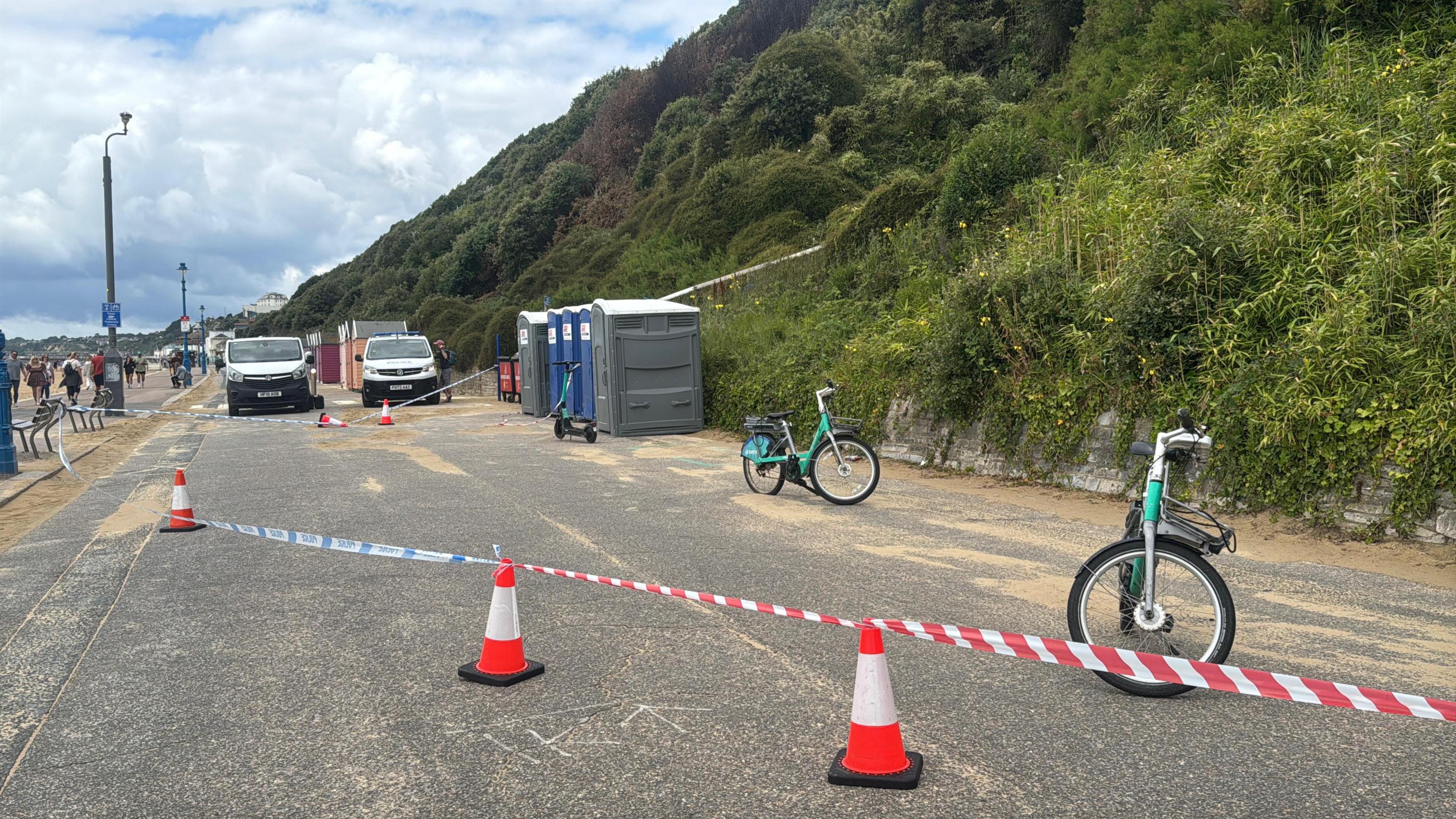 Five blue and grey portable toilets standing on a road next to a bush-covered cliff, with police tape tied to traffic cones around them, blocking the road