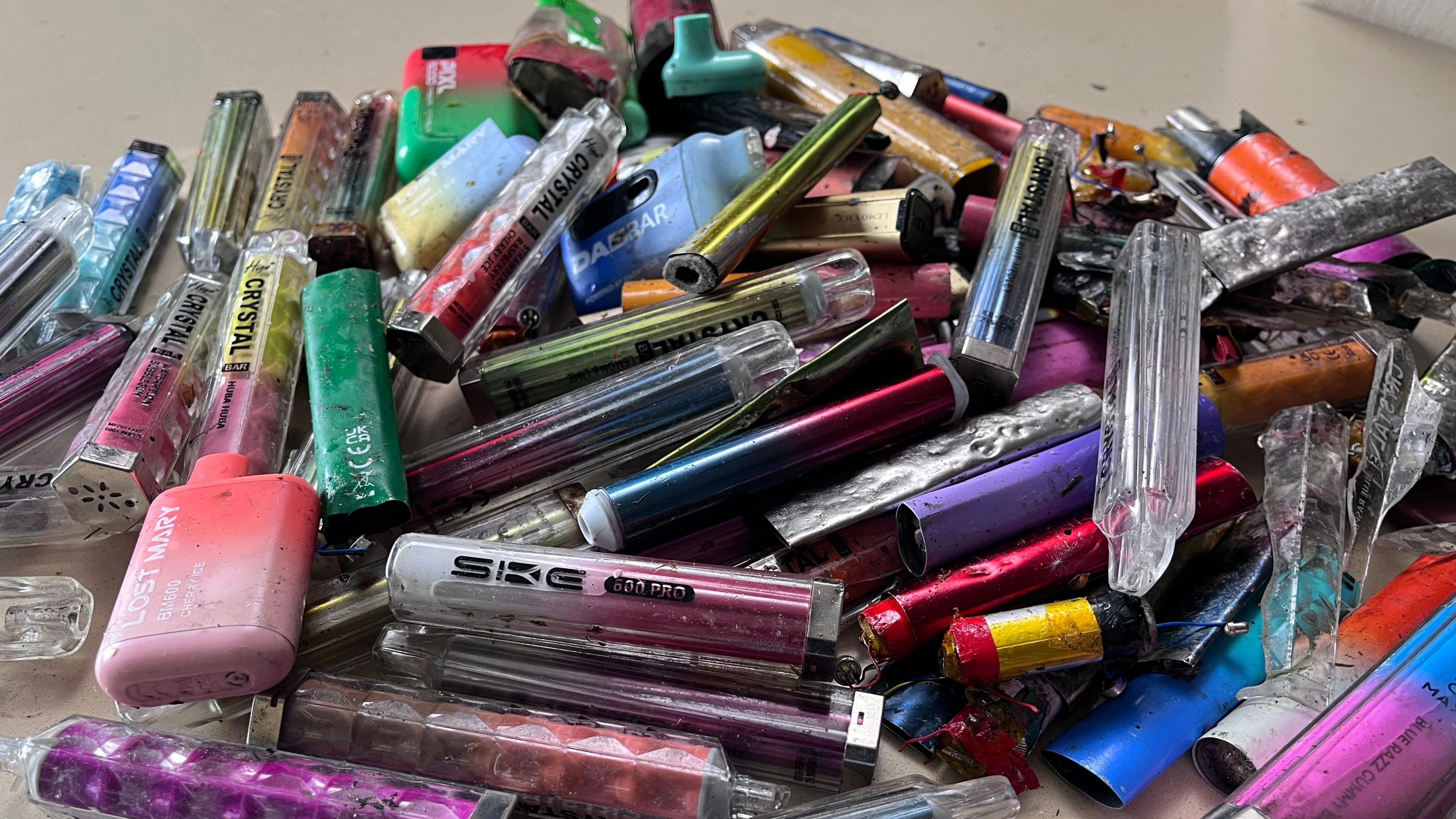 A pile of brightly coloured and different shaped vapes. They are in varying states but are all clearly used.