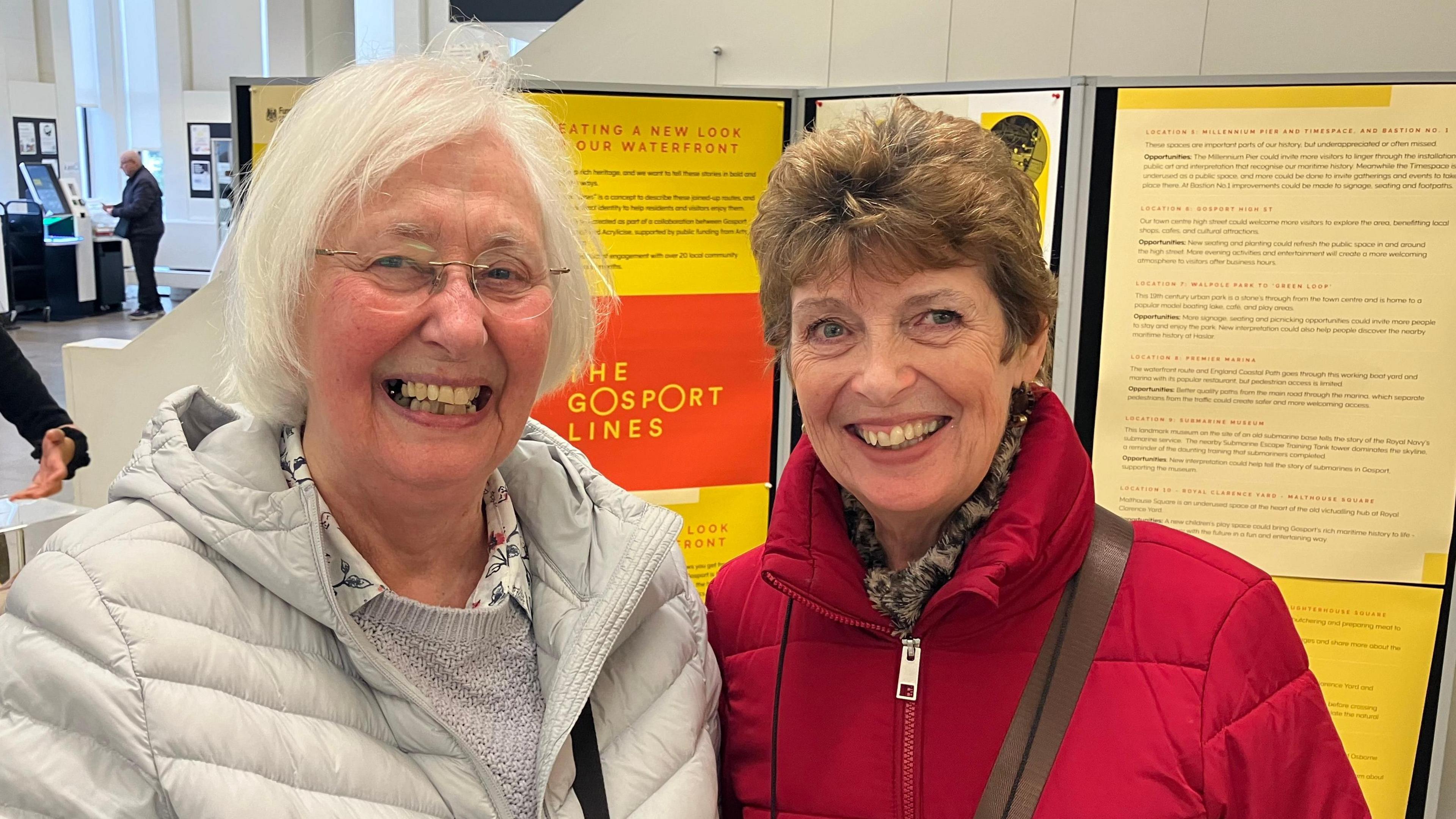 Two ladies stand in front of an information board about Gosport waterfront. One has white hair, glasses and a white coat and the other dark hair and a red coat.