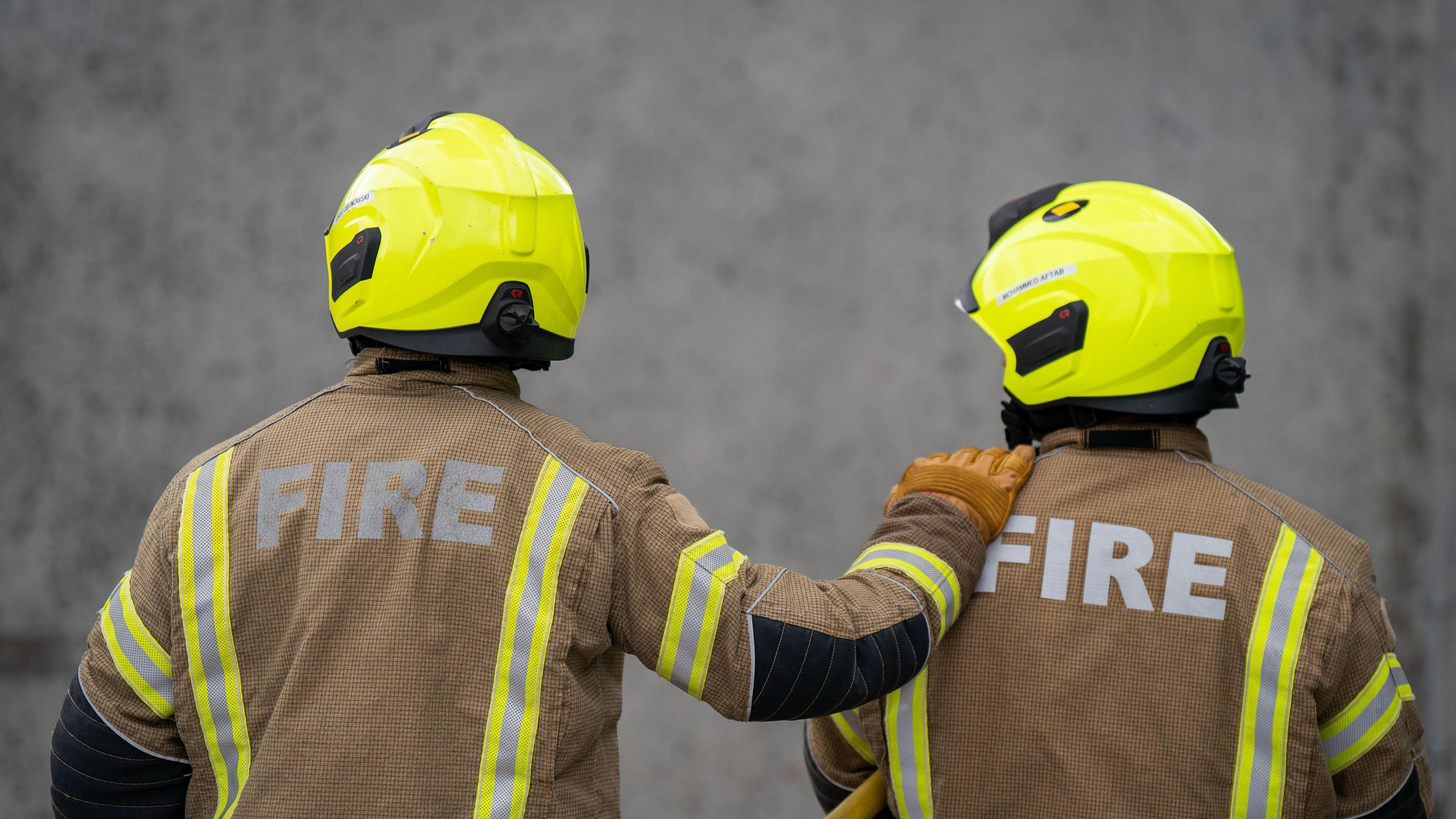 Stock image of two Firefighters in uniform facing away from the camera. The officer on the left has put his right arm on the other officers left shoulder.