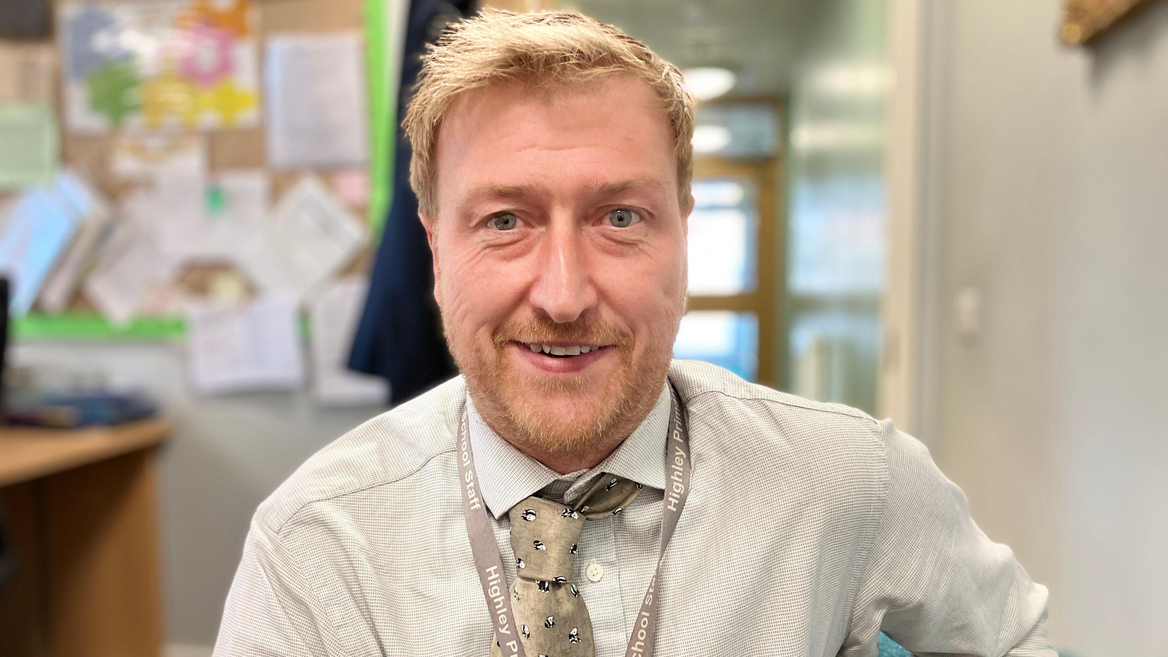 A fair-haired man man with stubble, wearing a cream shirt and tie, smiling at the camera inside an office with notes and paper pinned to the wall behind him
