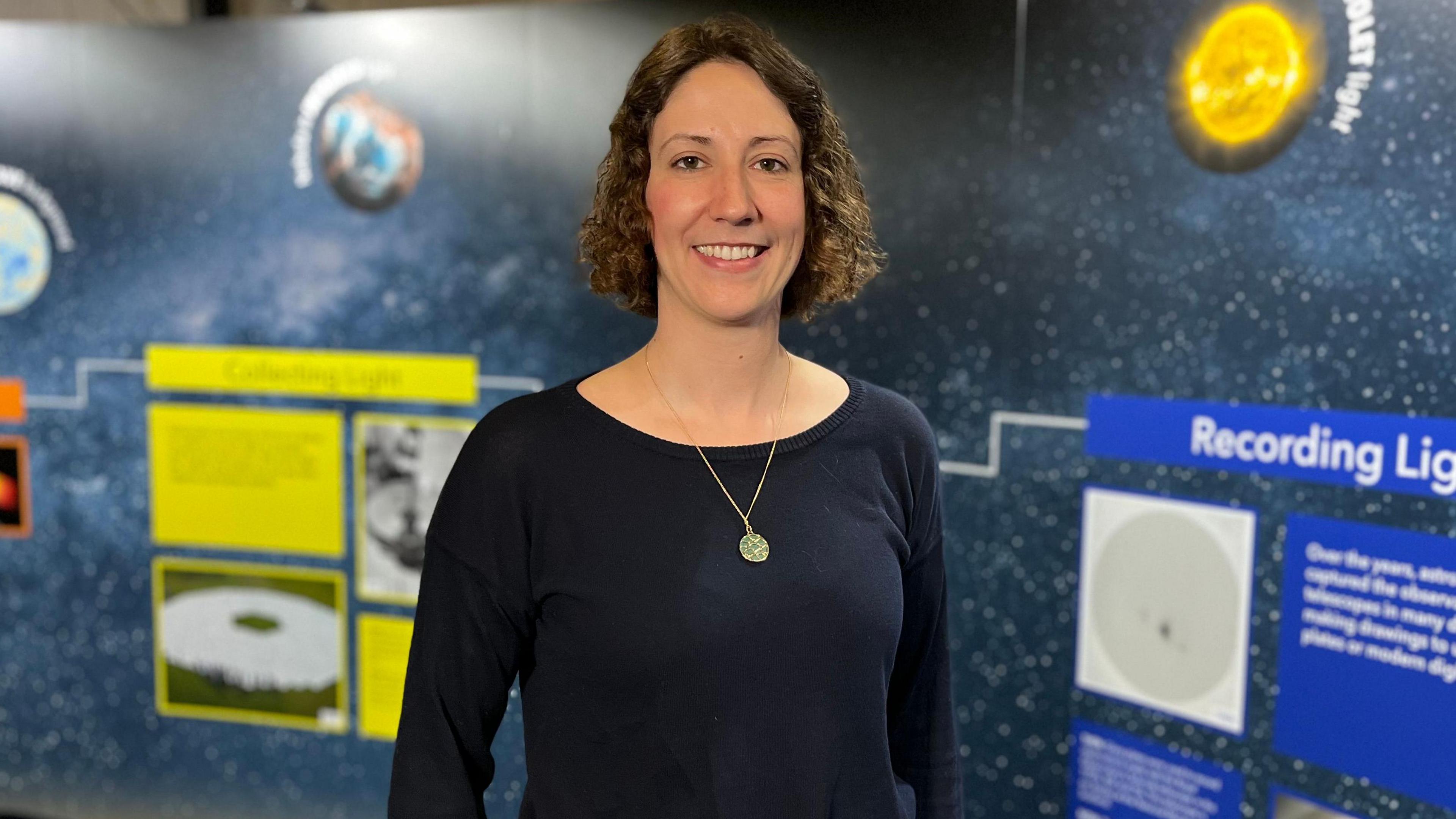 Cyrielle Opitom, a woman with short curly brown hair wearing a black boatneck top with a gold pendant necklace. She is smiling standing in front of a space backdrop.