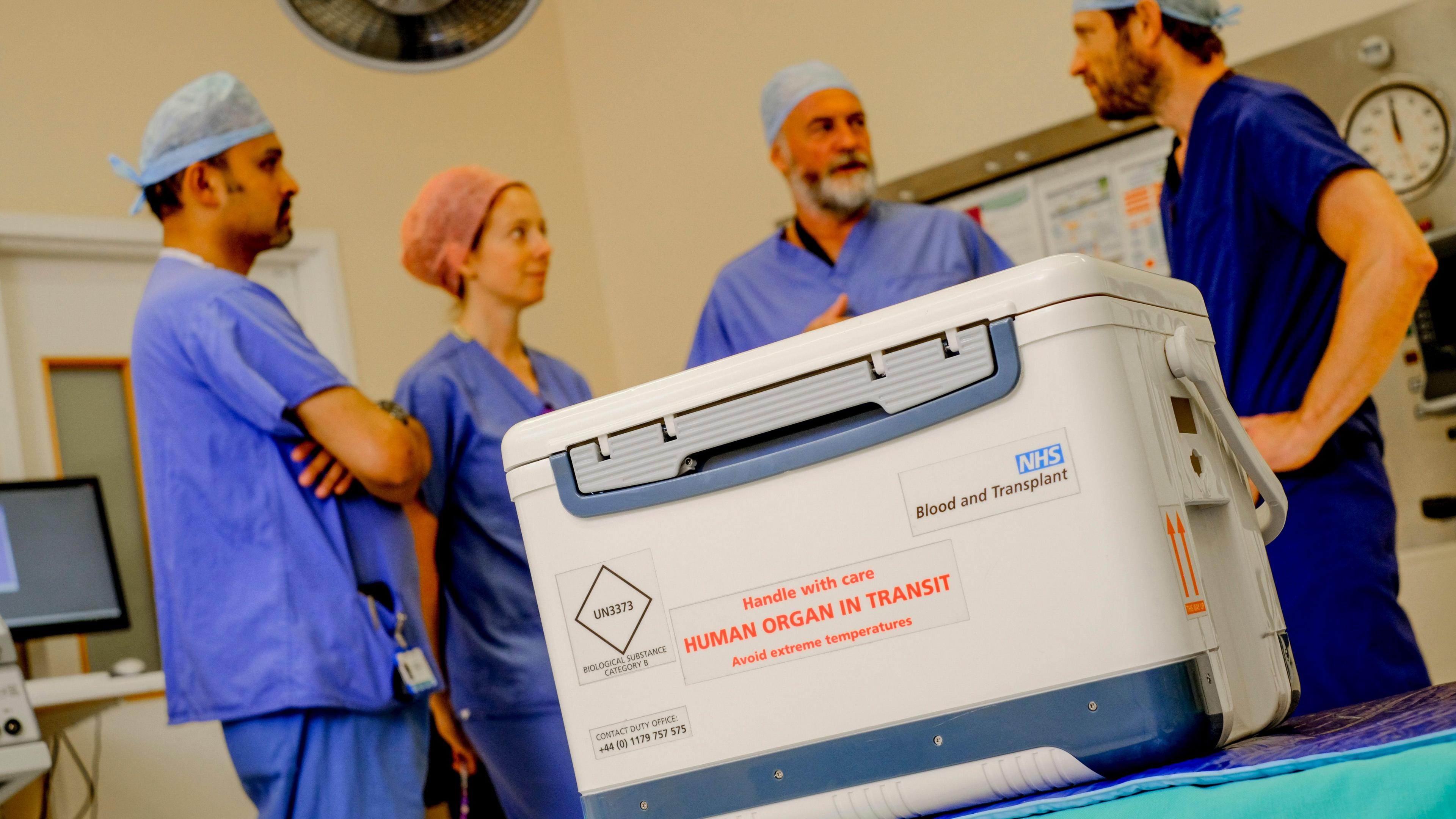Organ transplant box which says "human organ in transit". There are four doctors in the background in scrubs 