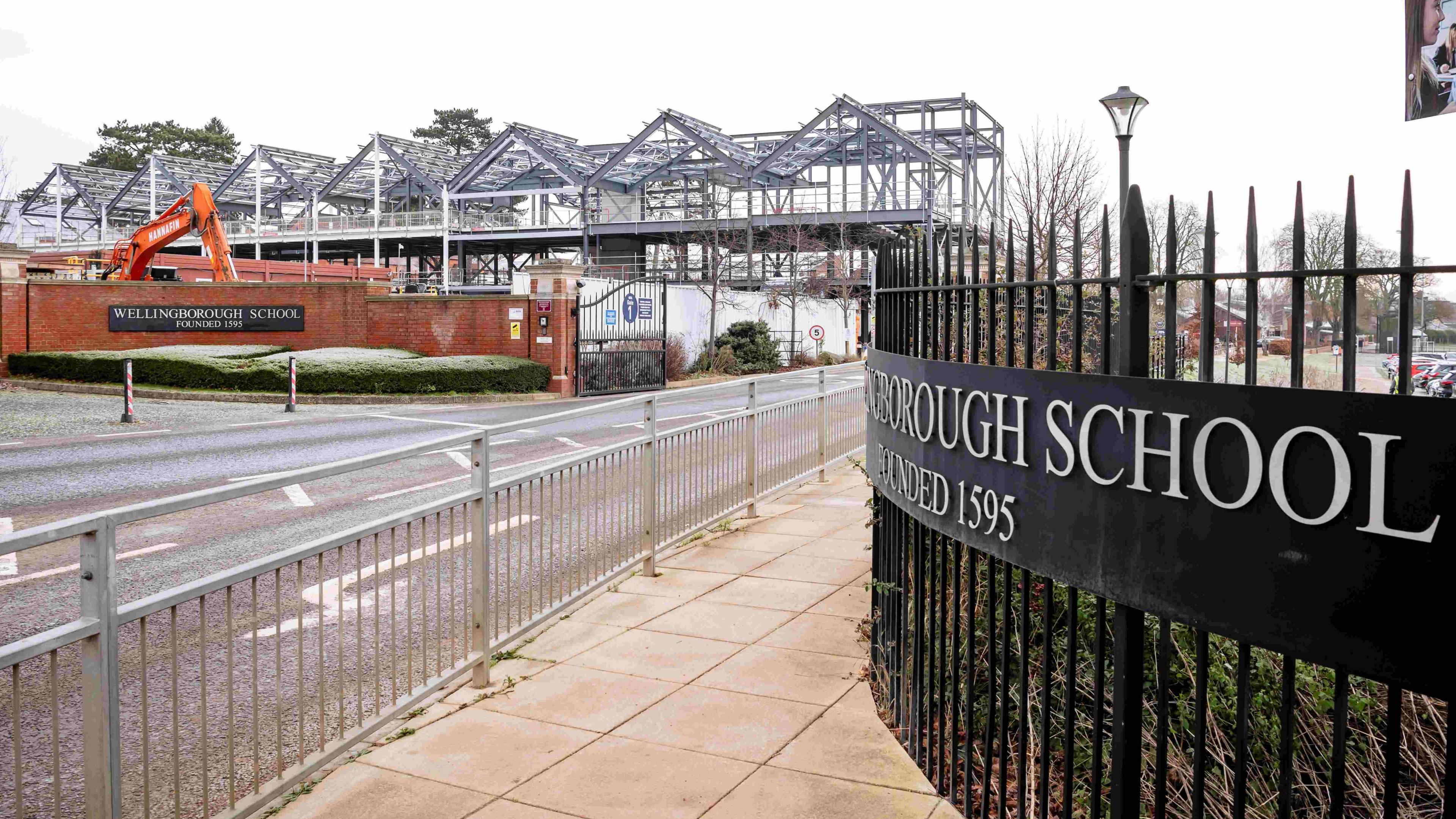 An entrance to Wellingborough School, showing a steel frame of the new school building being built. On a black metal gate reads "Wellingborough School, founded 1595".