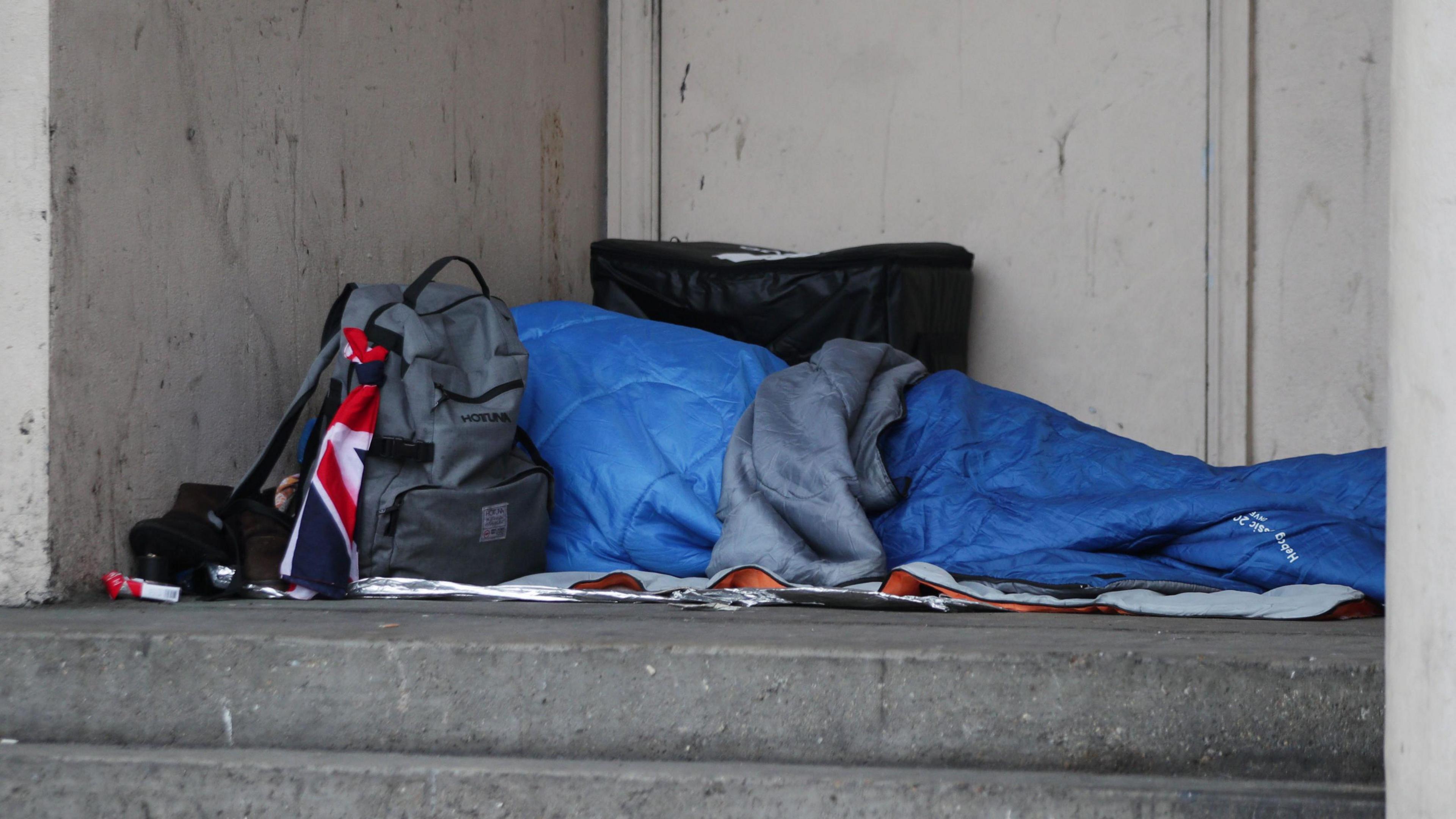 A rough sleeper in a blue sleeping bag on the ground with his grey back next to it 