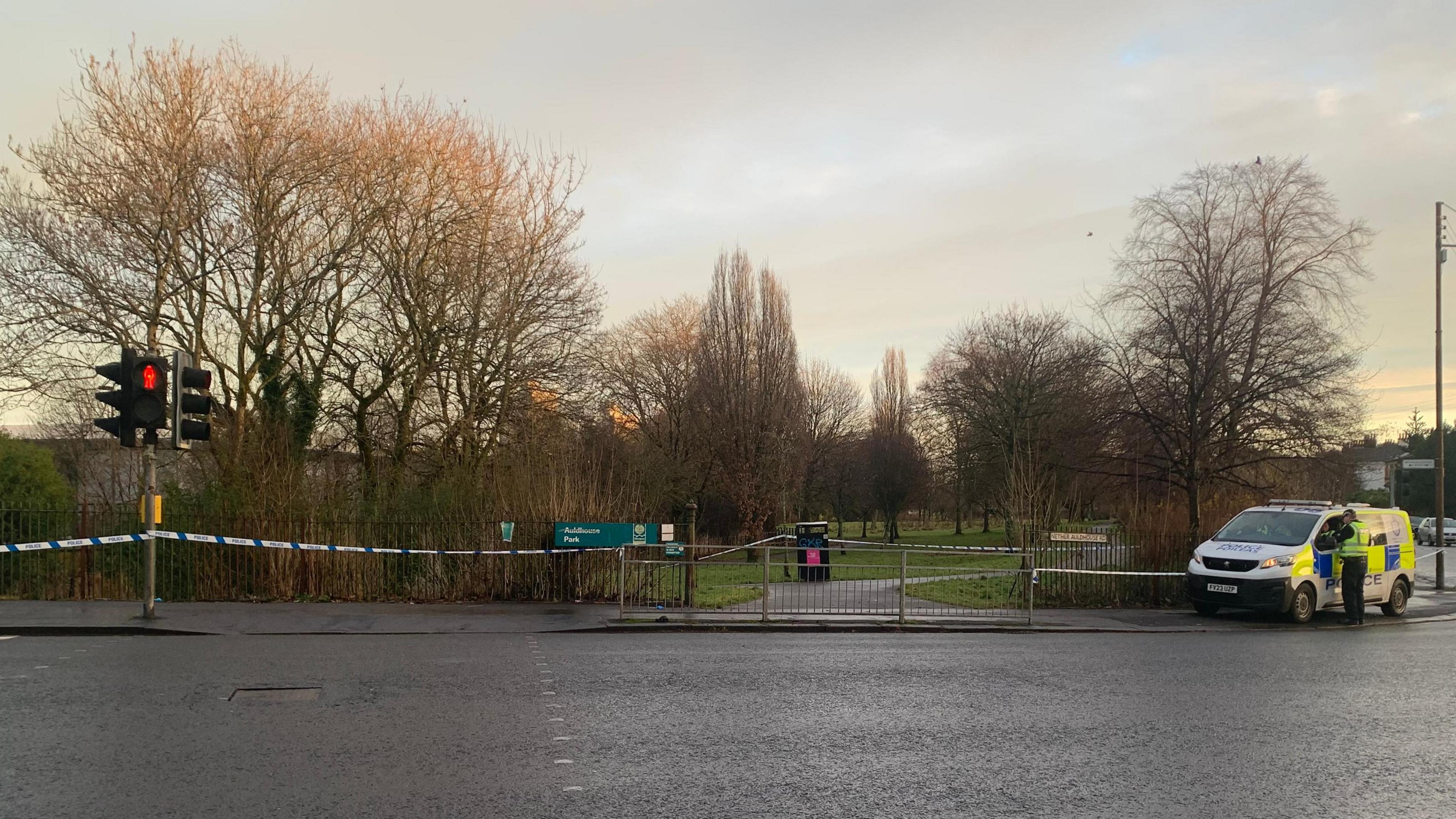 Entrance gate of Auldhouse park from a distance with police tape sealing off the area and a Police Scotland van parked on the pavement at the street corner