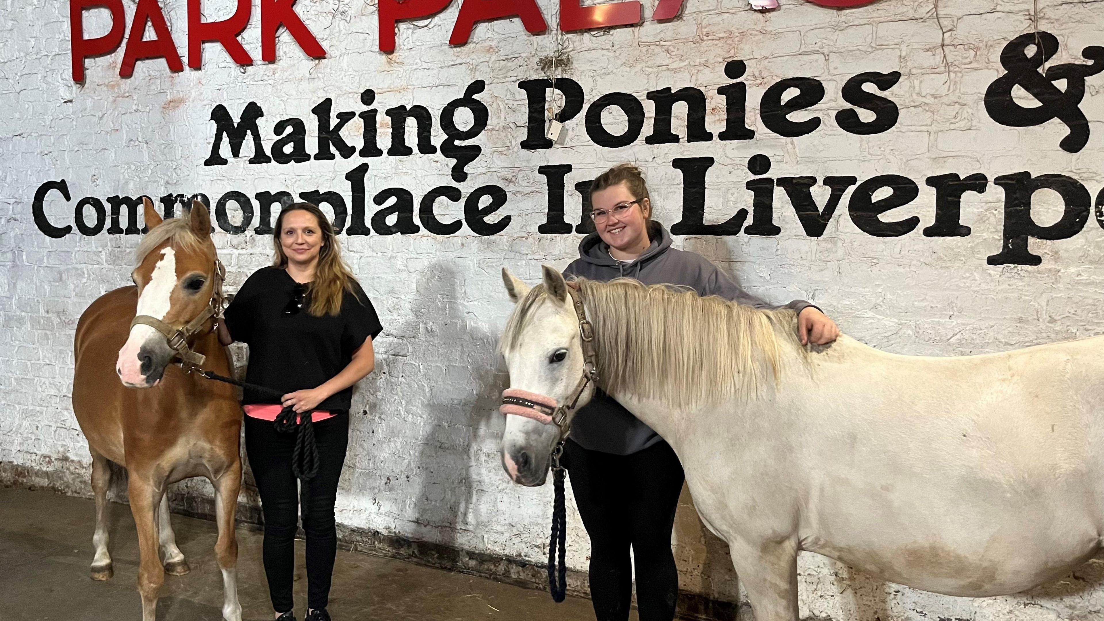 Pam Allen, who has long blonde hair, stands next to a light brown pony with a white patch on its face, while Megan Turley, who has dark blonde hair tied back, poses with her arm around a white pony