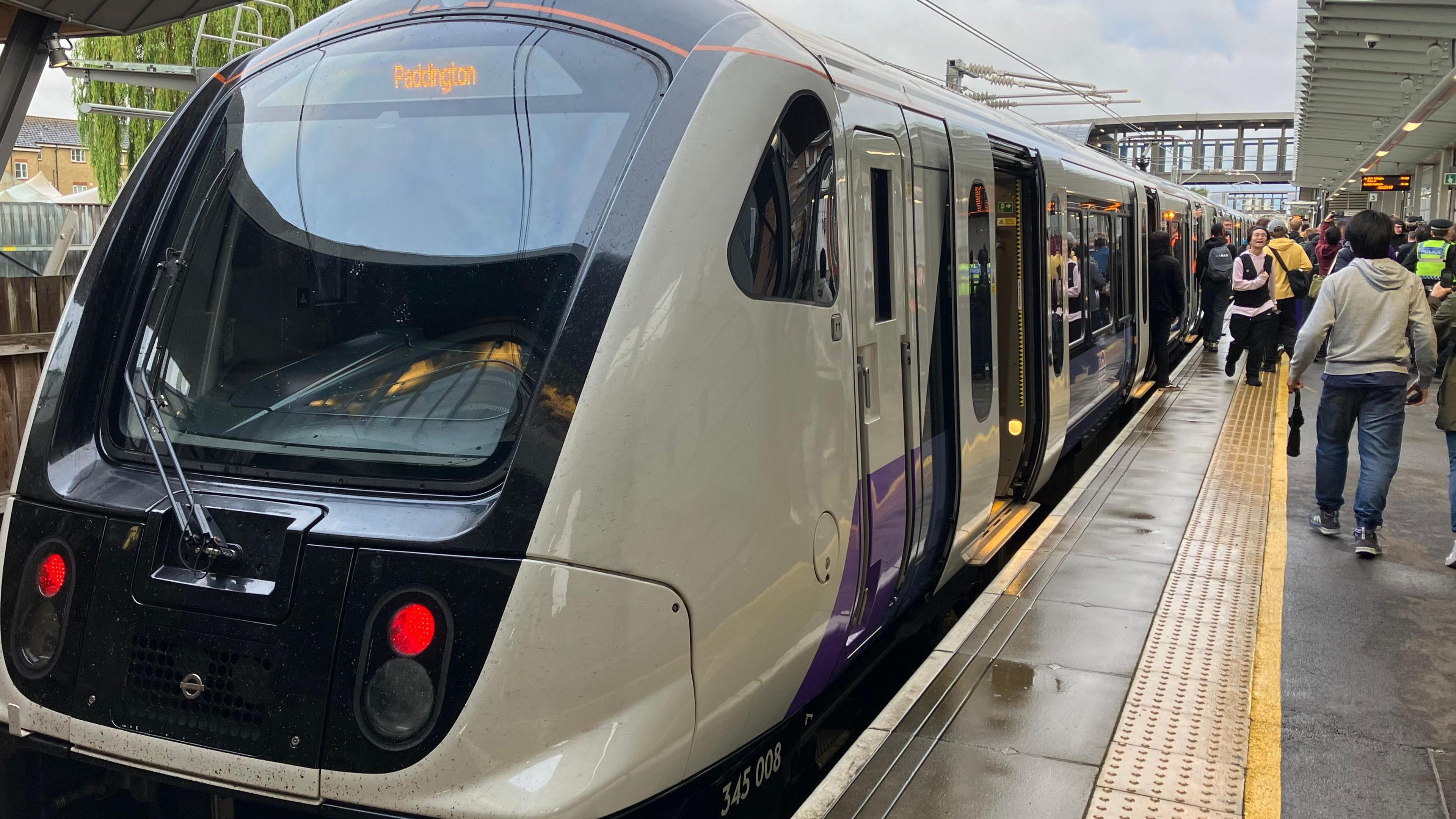 An Elizabeth line train at the platform with its doors open. There are people walking along the platform beside the train. 