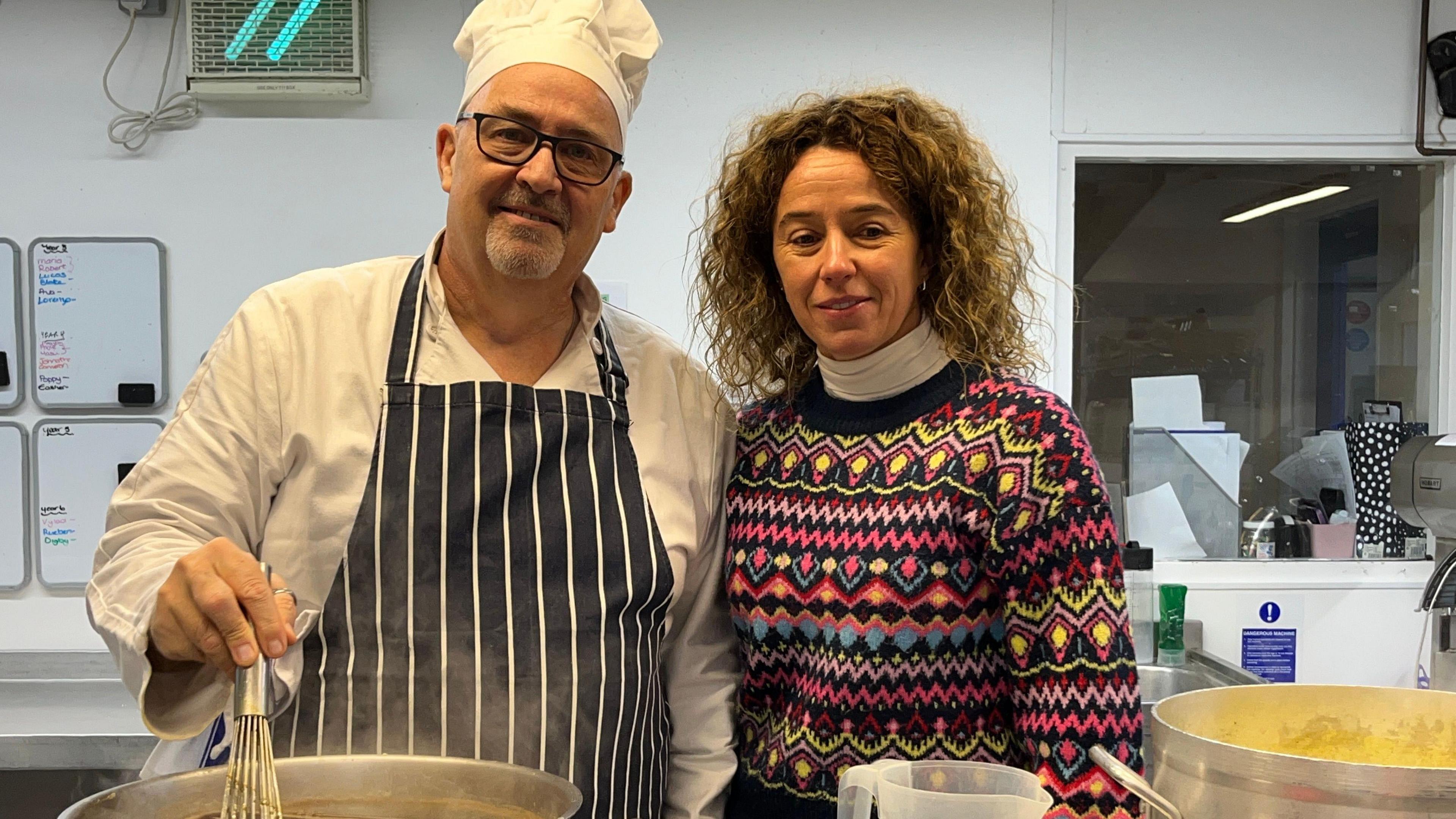 A man in chef's whites and a blue apron stirs a pot of gravy, helped by his wife.