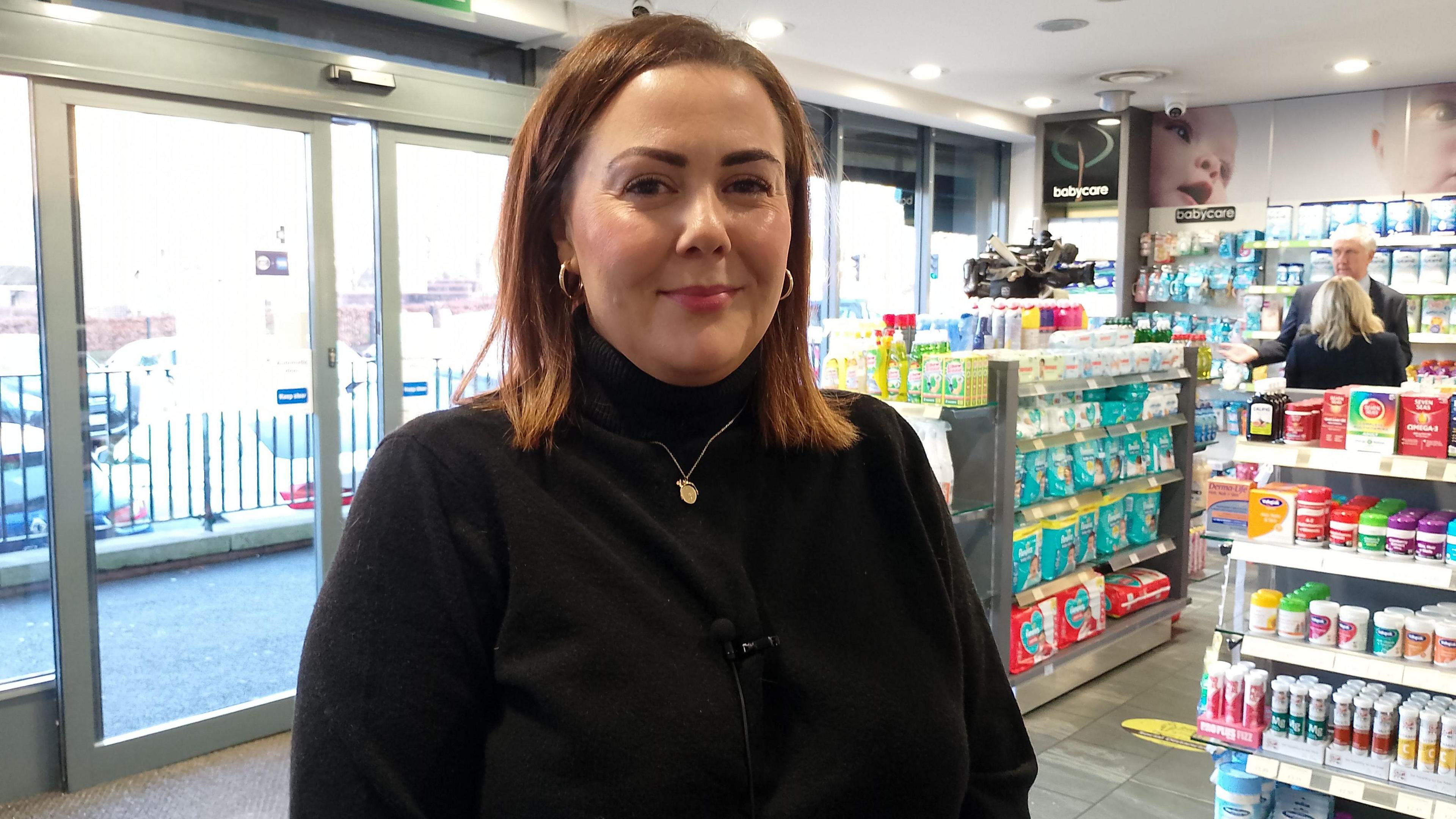 Rachel Spiers stands in a pharmacy smiling. She has shoulder length auburn hair and brown eyes. She is wearing a black cardigan and gold chain.