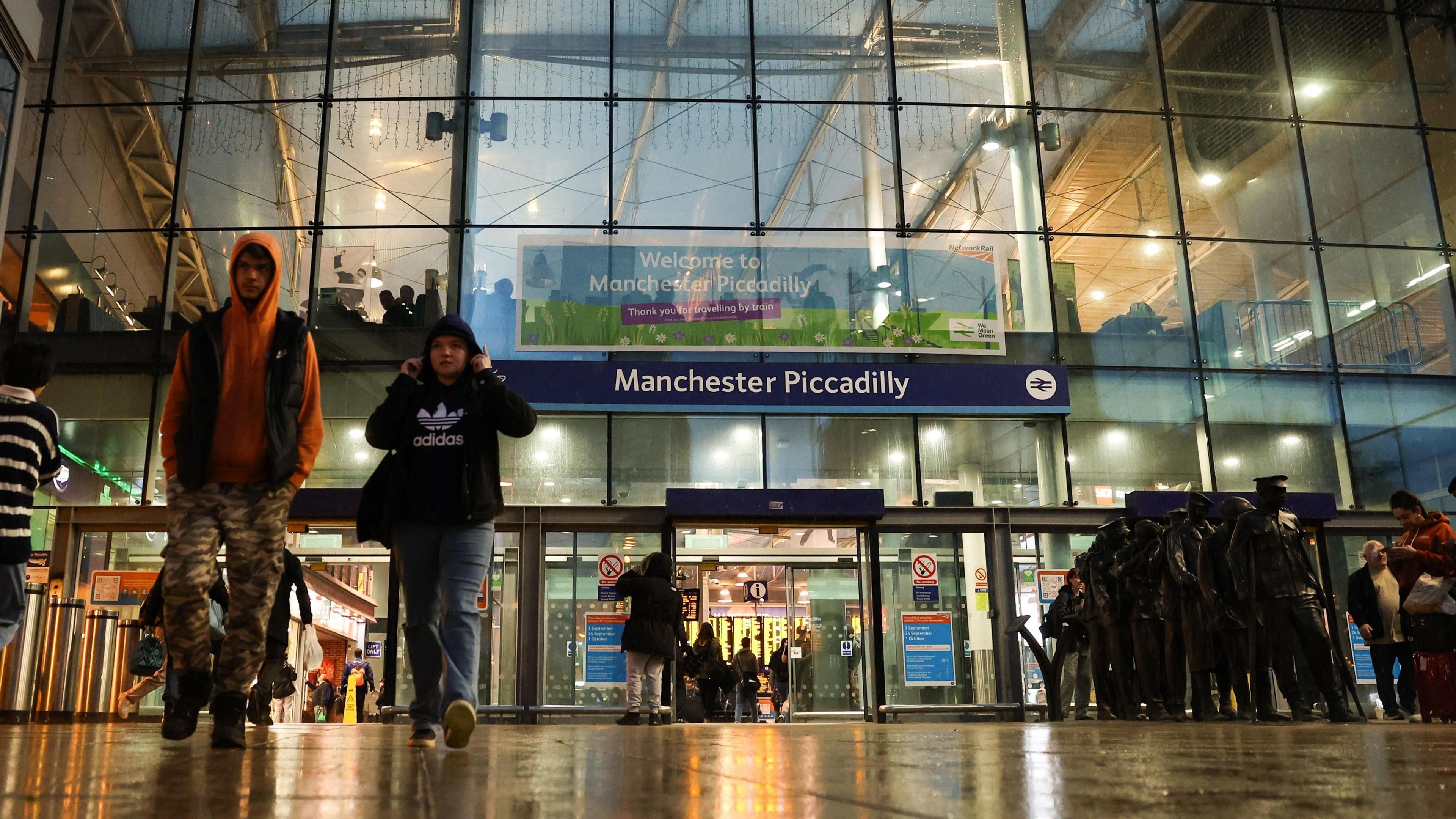 People walk out of the glass-fronted entrance to Manchester Piccadilly train station on a wet day.