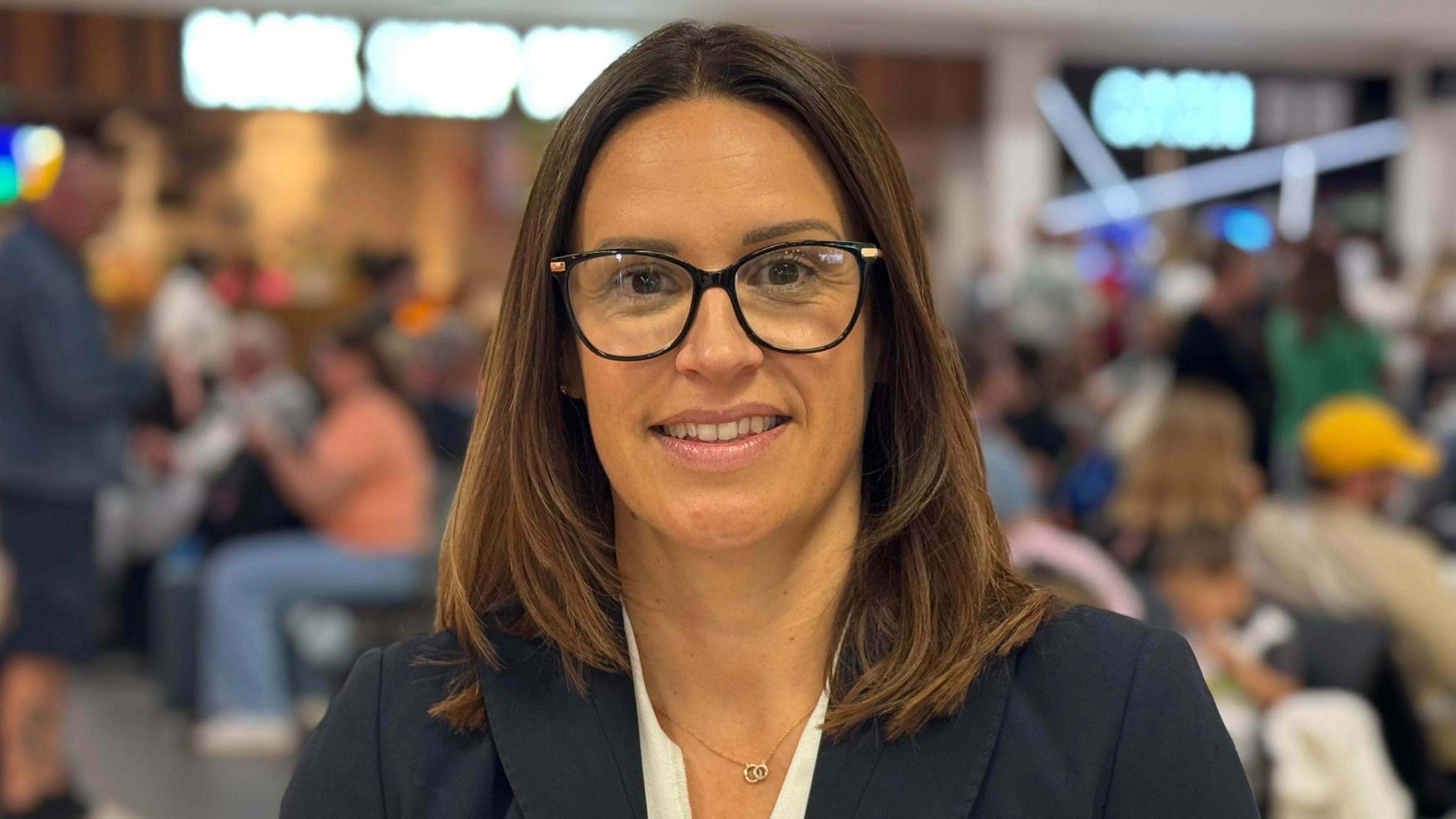 Clare Armstrong is smiling at the camera. She is wearing dark rimmed glasses, a blue blazer and has shoulder length dark hair. She is standing in the departure lounge with anonymous passengers sitting in the background awaiting flights.