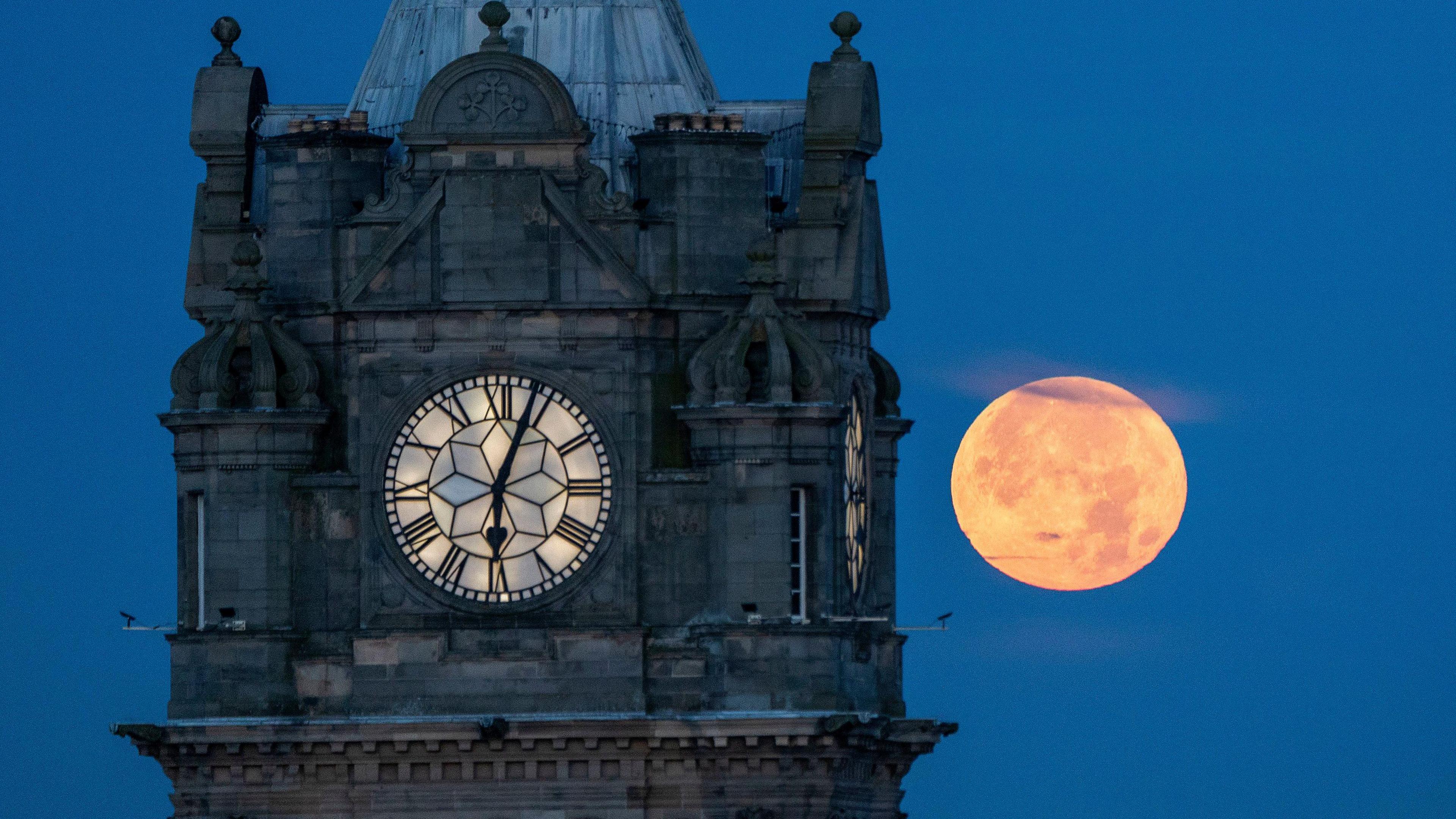 Clock face in foreground with a large, bright orange-coloured moon against a dark blue sky in the background