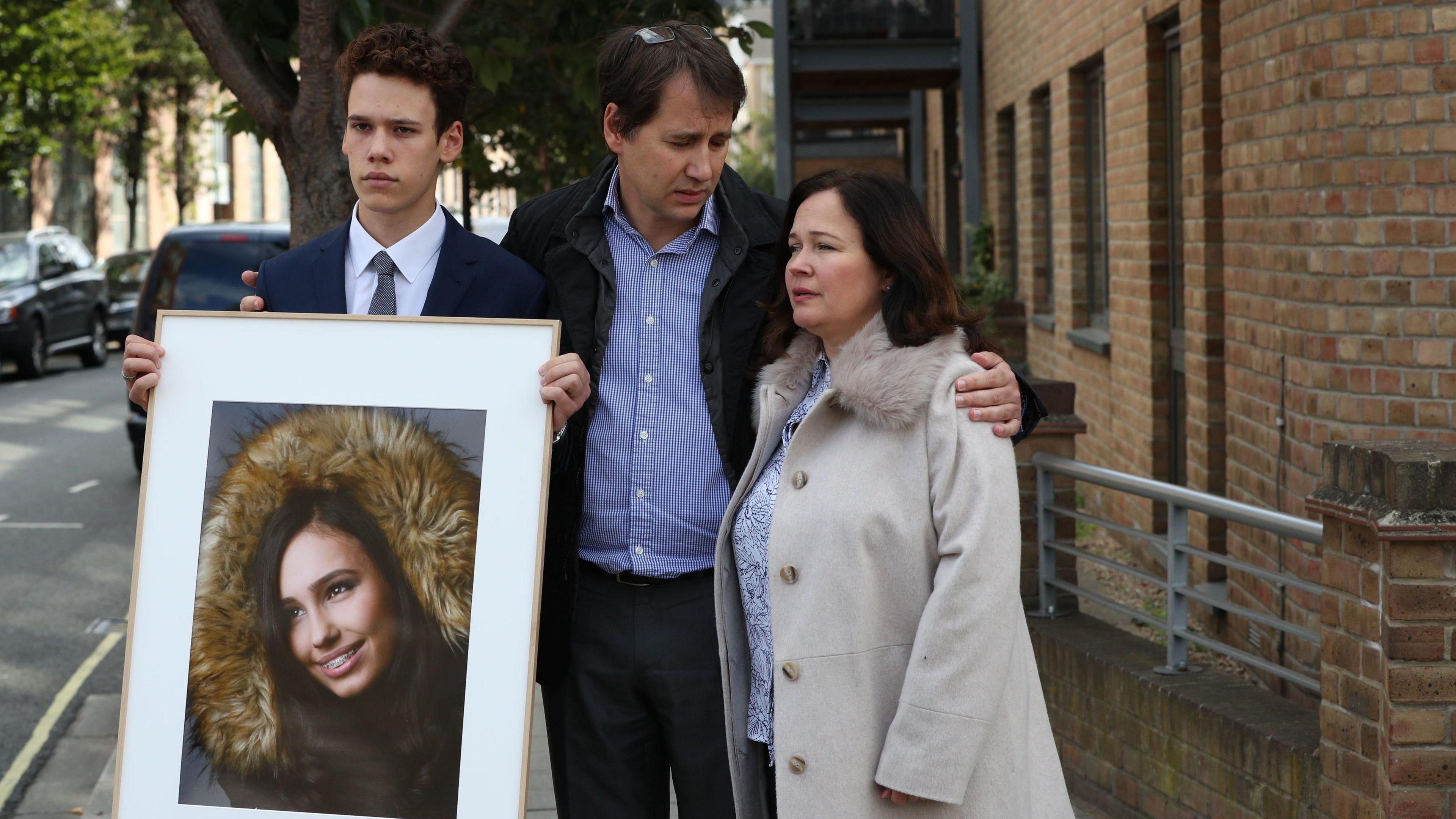 A family is holding a framed image of Natasha. Natasha has brown hair and is wearing a fur hood. Her father is wearing a blue shirt and has one arm around Natasha's mum who is wearing a long cream jacket. Their son Alex is dressed in a suit. He is holding the picture of Natasha.