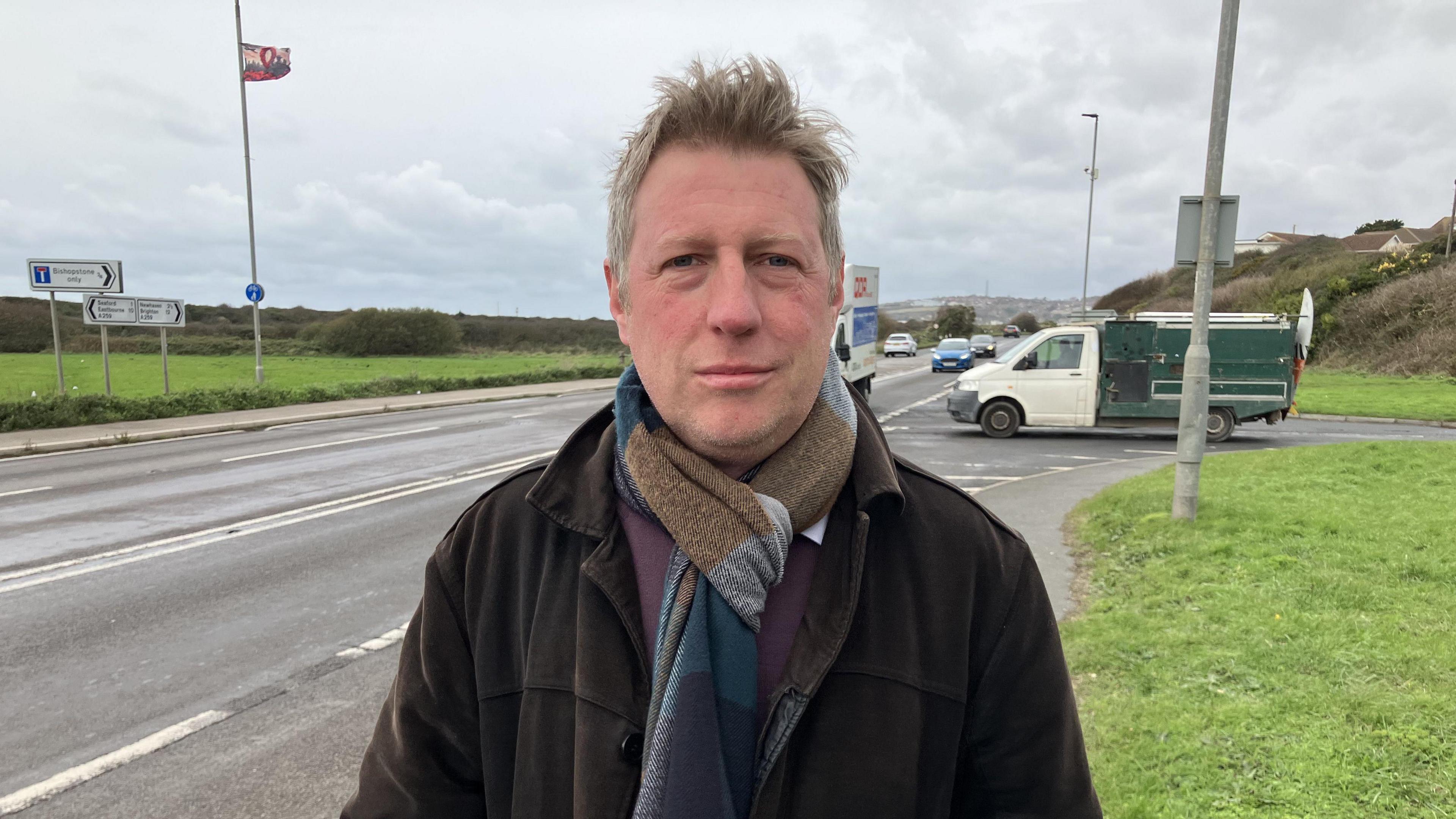 Member of Parliament James MacCleary standing by the side of the A259 road on the pavement. He has his hands in his pockets and he is wearing a brown jacket and striped tie. Cars and a van can be seen driving on the road behind him. It is a grey, cloudy day. 