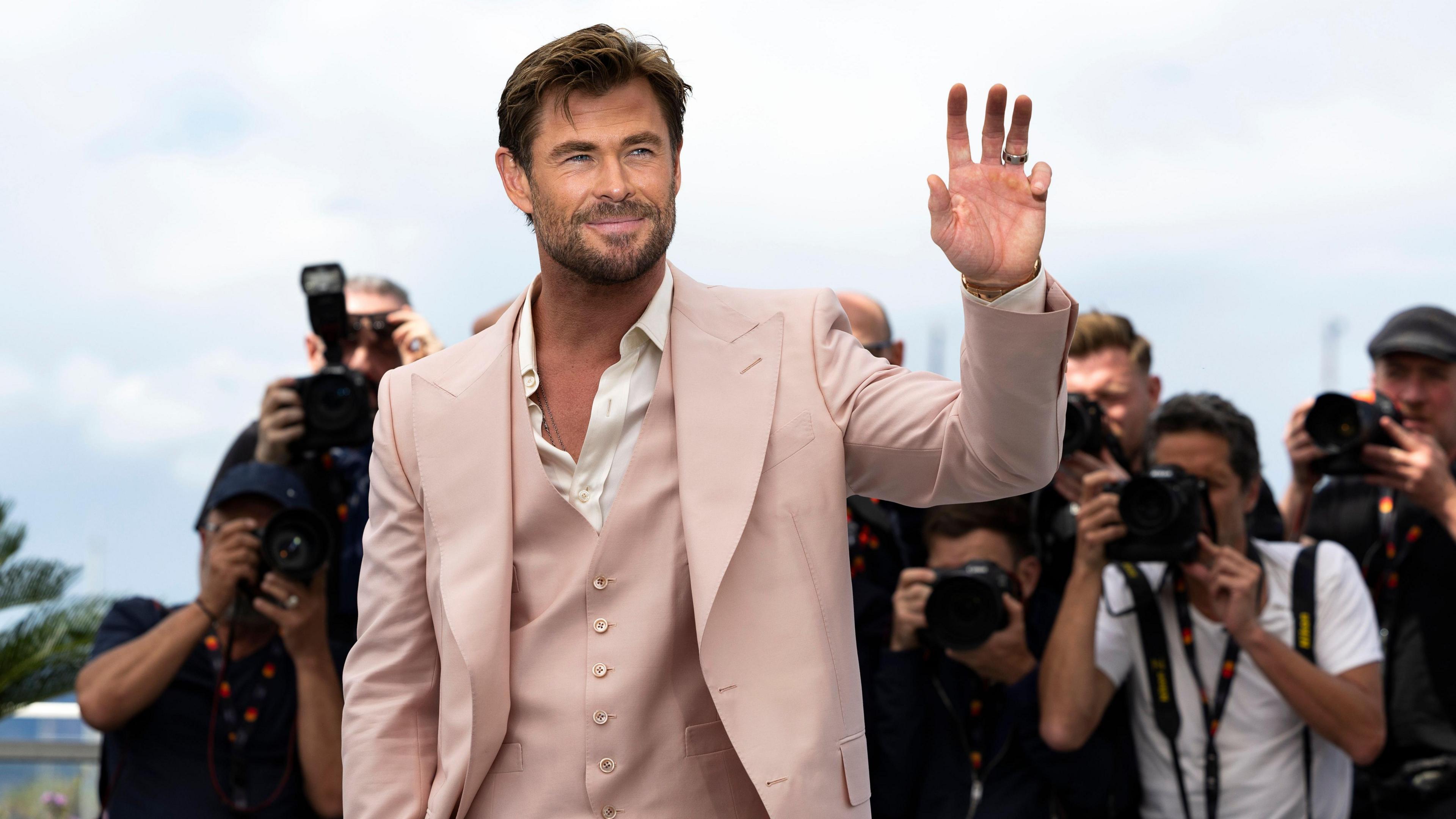 Chris Hemsworth attends the photocall for 'Furiosa: A Mad Max Saga' during the 77th annual Cannes Film Festival, in Cannes, France. He waves at the crowd wearing a salmon-pink blazer, waistcoat and white shirt. A crowd of photographers stand behind him.
