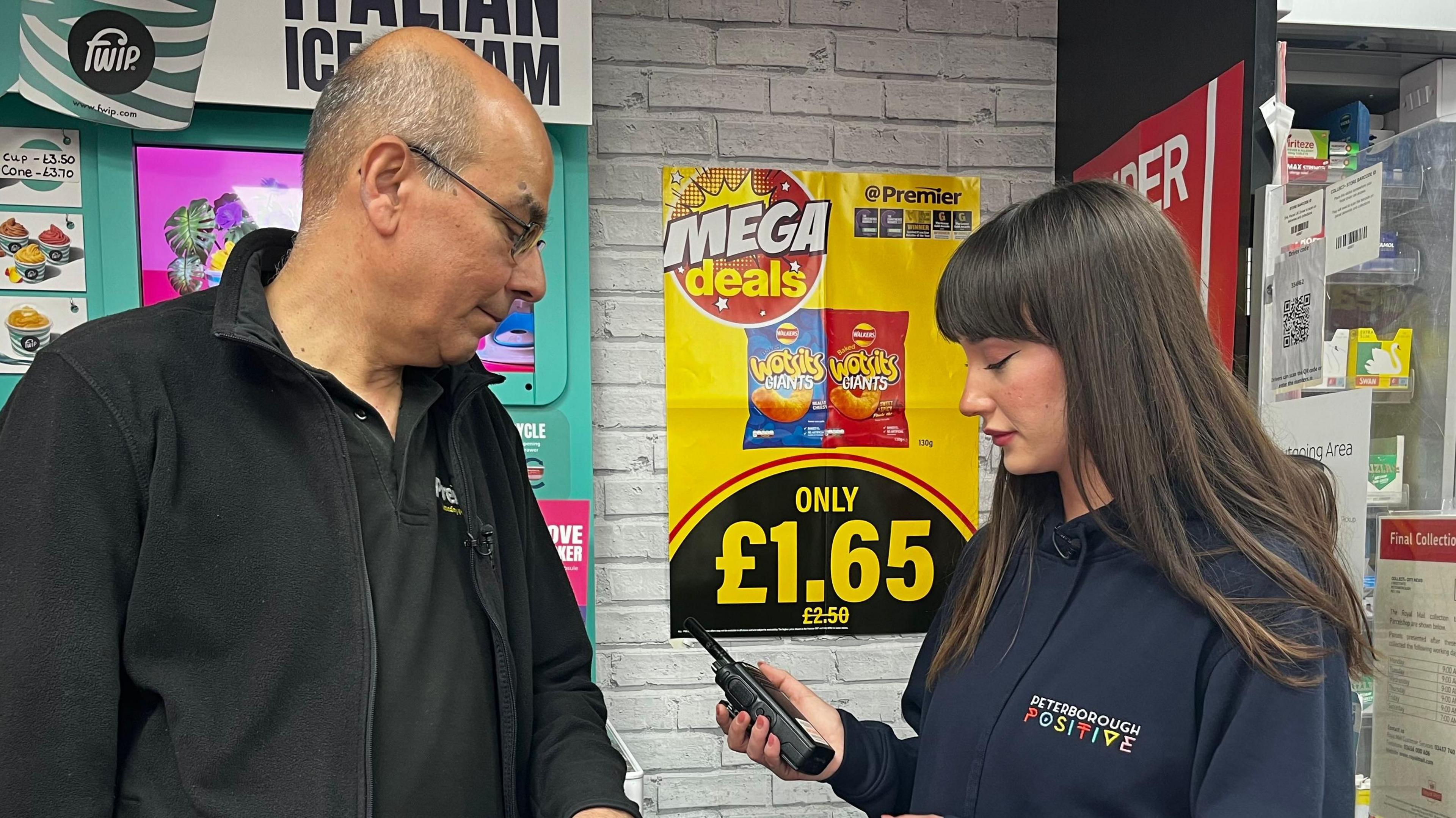 Vidyut Soni is being shown a radio by a woman in a hooded top with "Peterborough Positive" written on it. They are standing in a shop with a poster behind that reads: "Mega deals only £1.65". 