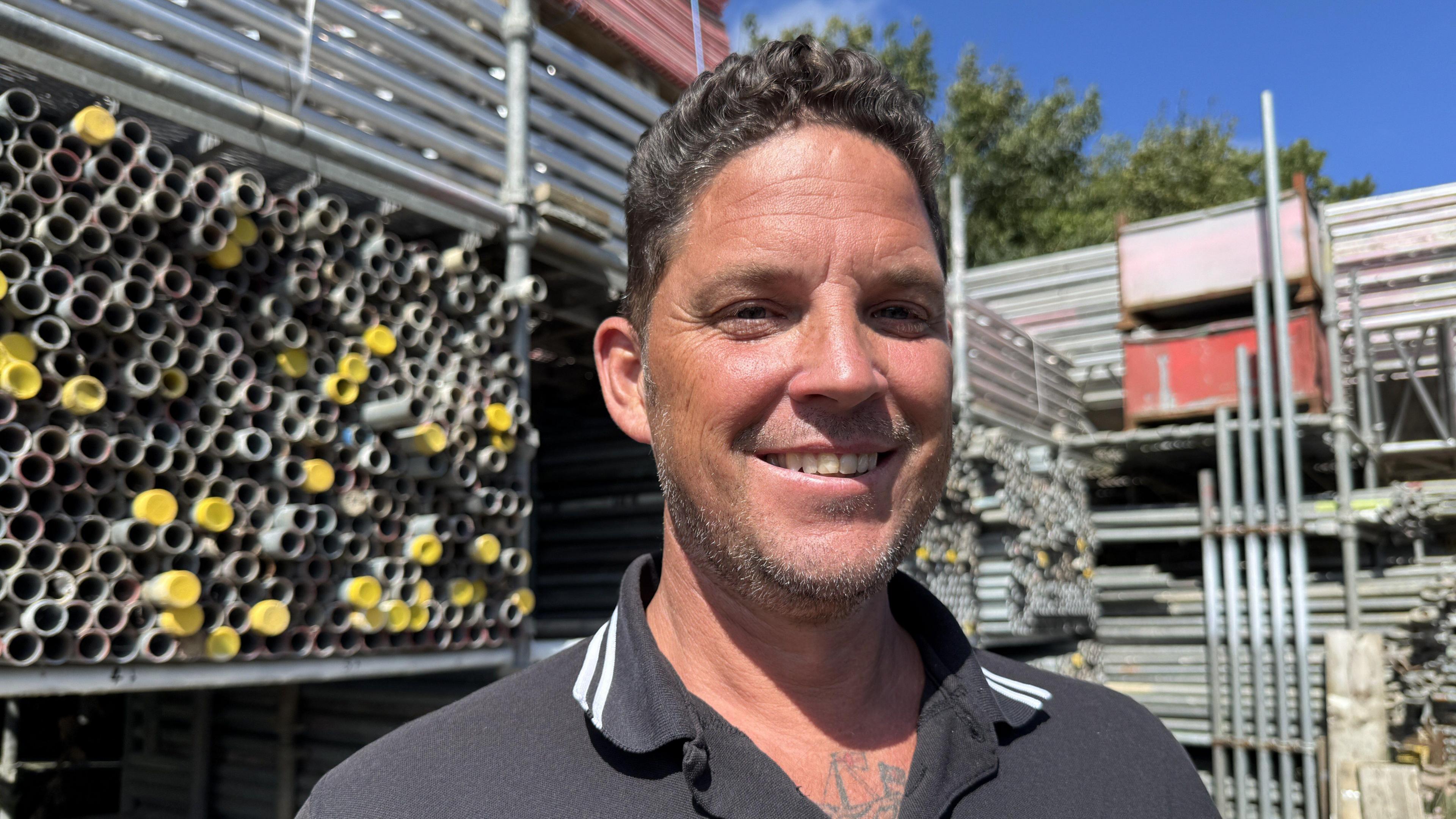 A man stands in front of a neatly arranged storage area containing scaffolding equipment and metal pipes. He is wearing a black polo shirt featuring white collar trim and a visible "AA Scaffolding Solutions" logo on the chest. Behind him, the storage area displays stacks of metal pipes.