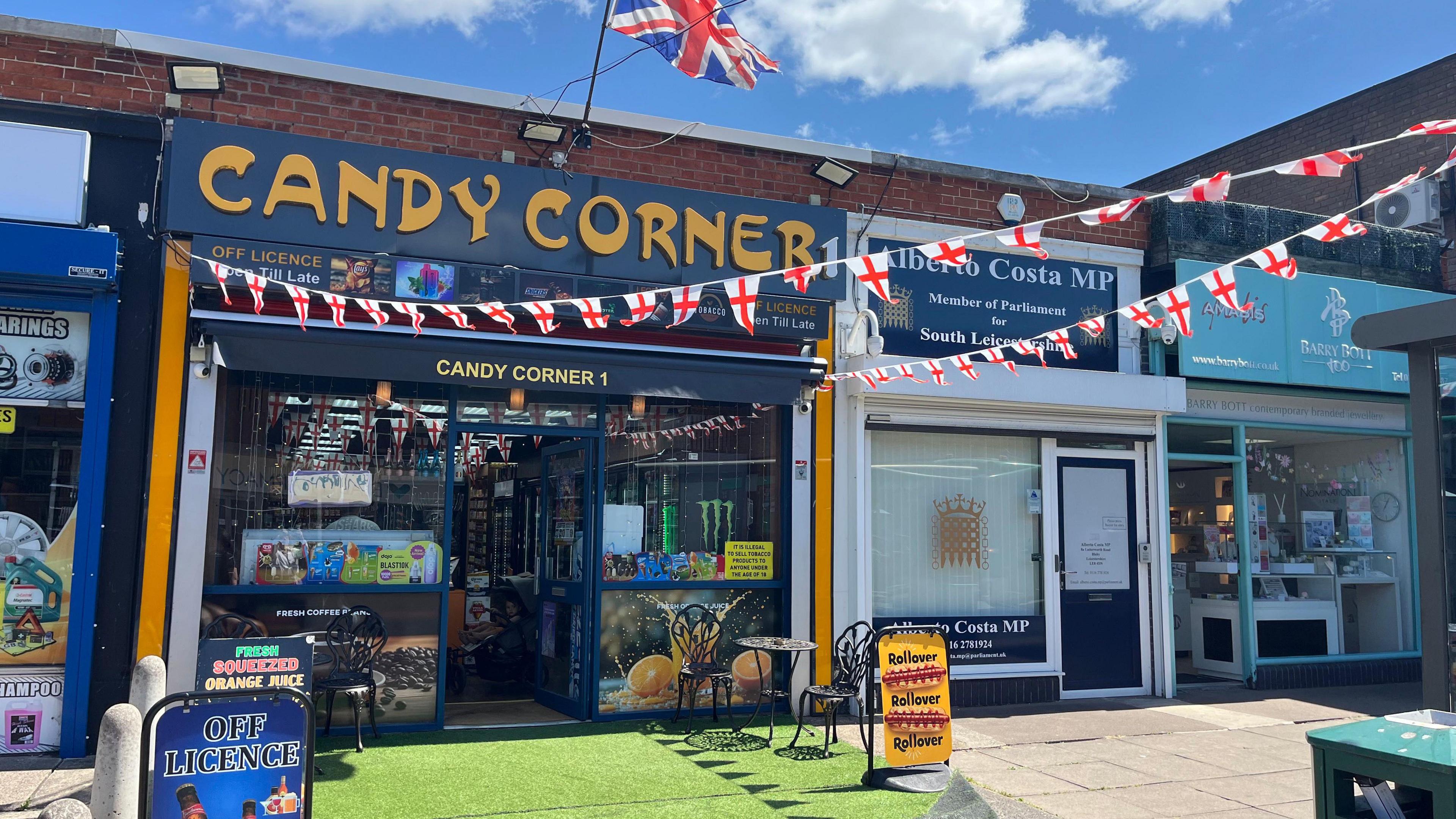 A shop called Candy Corner - with a red security shutter. Artificial grass matting is on the ground outside. There are St George's flags hung up either side of the shop front.
