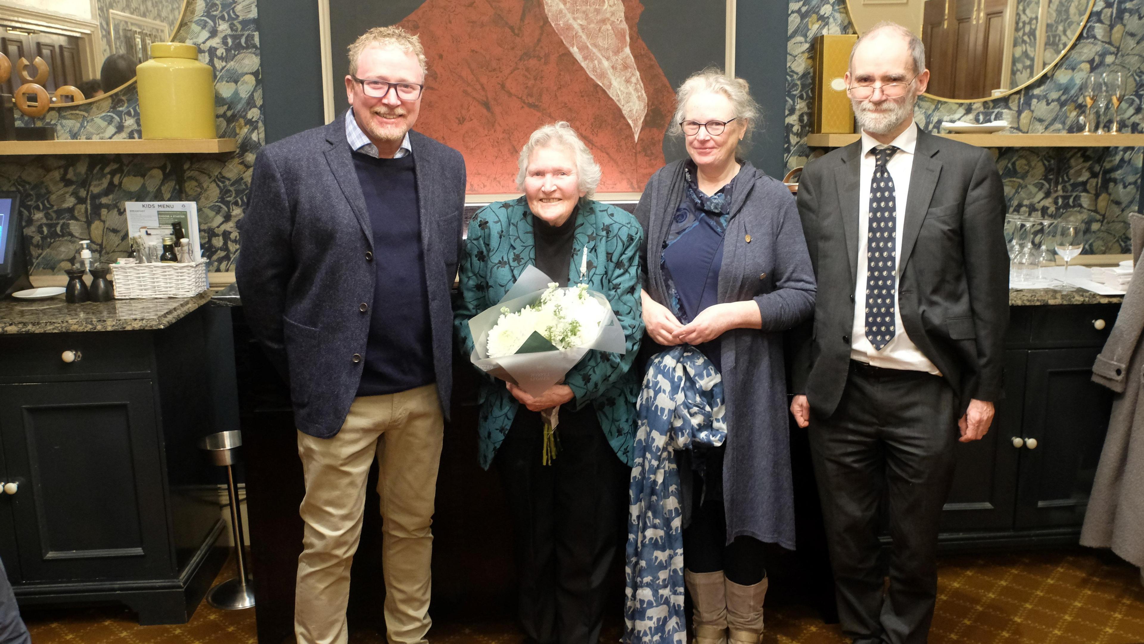 Gill Green standing in a line with three members of her family. From left-to-right there is John Green, her son, Gill Green, Imogen Allen, her daughter, and James Allen, her son-in-law. Mrs Green is holding a bouquet of white flowers.