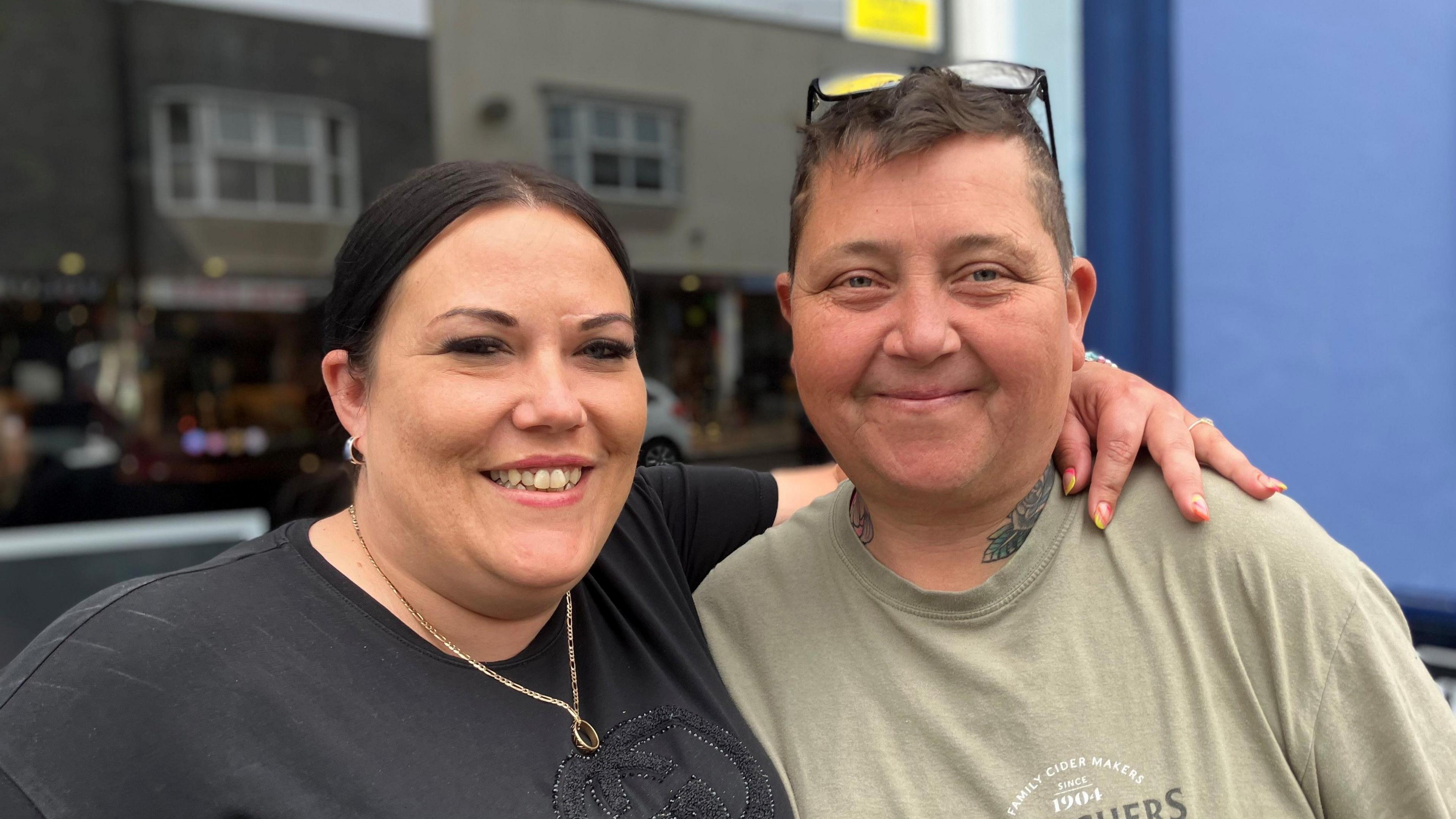 Sam Wright and Donna Shepherd are standing in the street with their arms around each other. They are smiling at the camera. One of the women has black hair and is wearing a black T-shirt while the other is wearing an olive green T-shirt and has a pair of glasses on the top of her head.