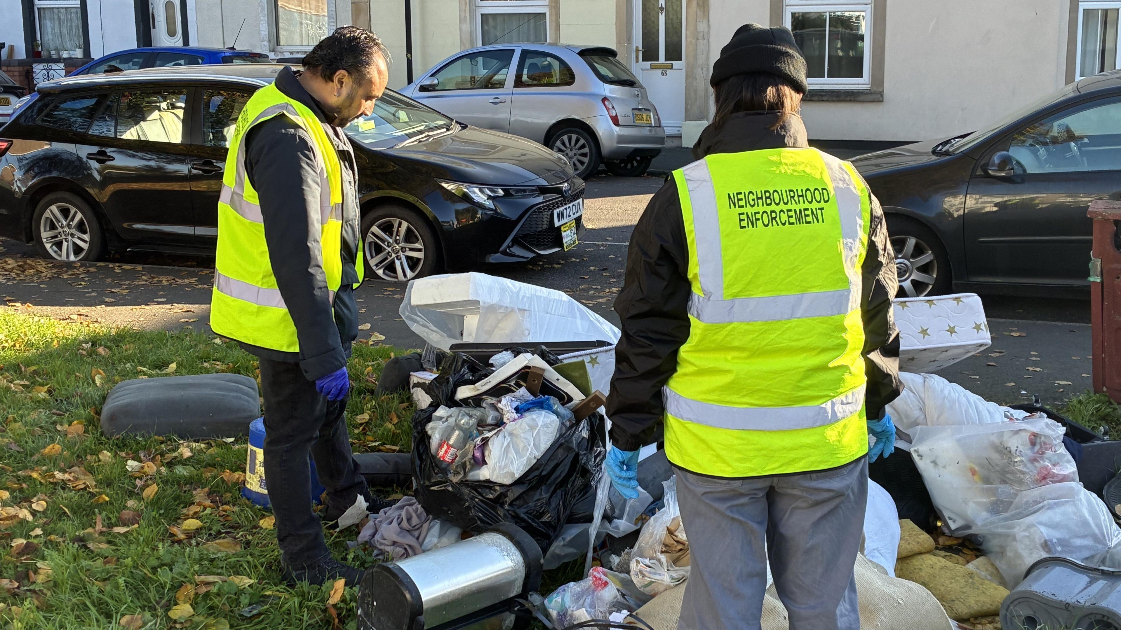 Two people wearing yellow high-vis jackets with the words 'neighbourhood enforcement' are looking down at a large pile of fly-tipped rubbish. They are standing next to a residential street which has lots of cars parked on the road. On the grass is a black bin bag with plastic waste inside, as well as household items like a plastic bin, mattress, pillow and rug.