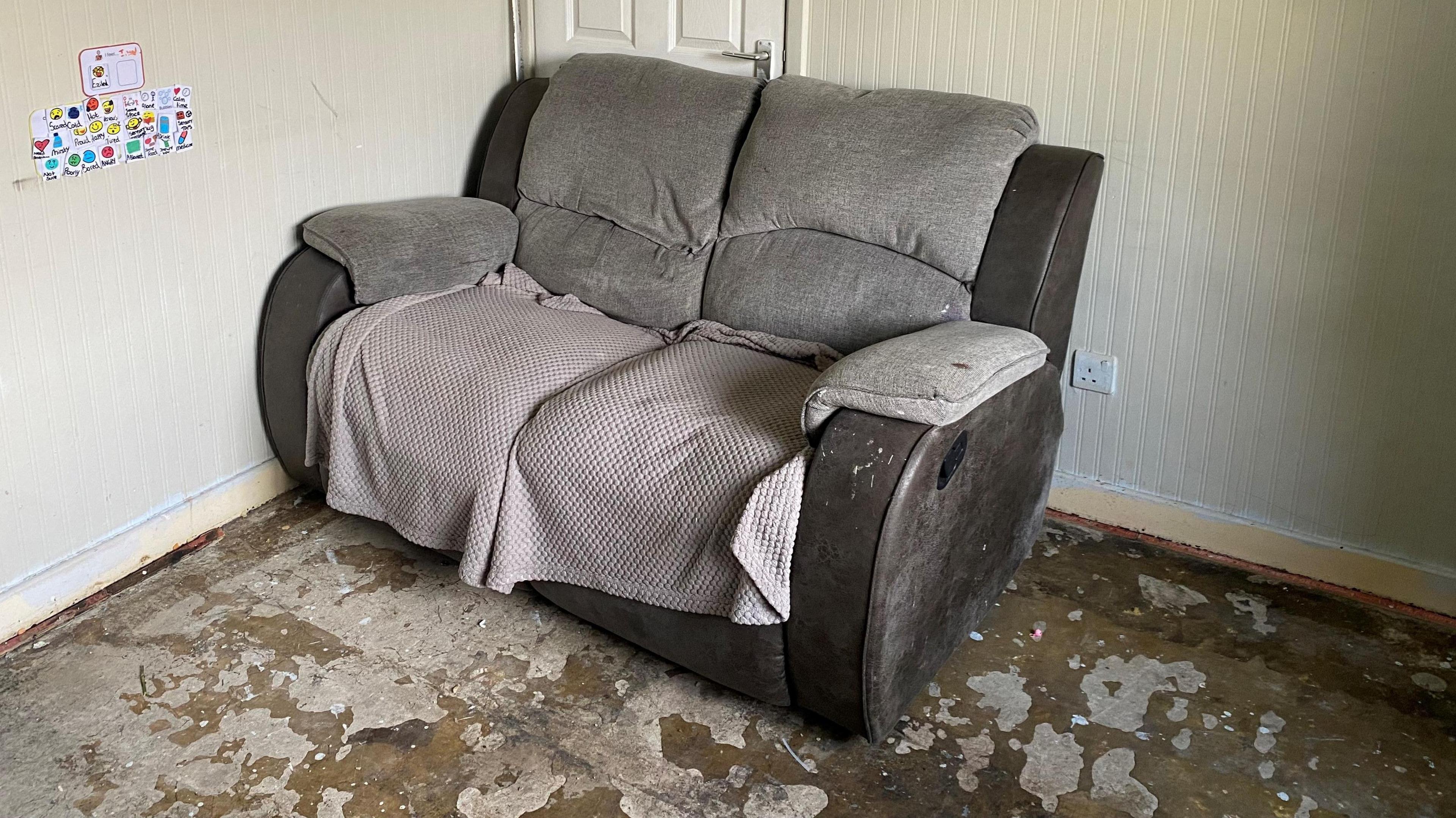 A brown two seater sofa covered with a grey blanket sits on a bare wooden floor.