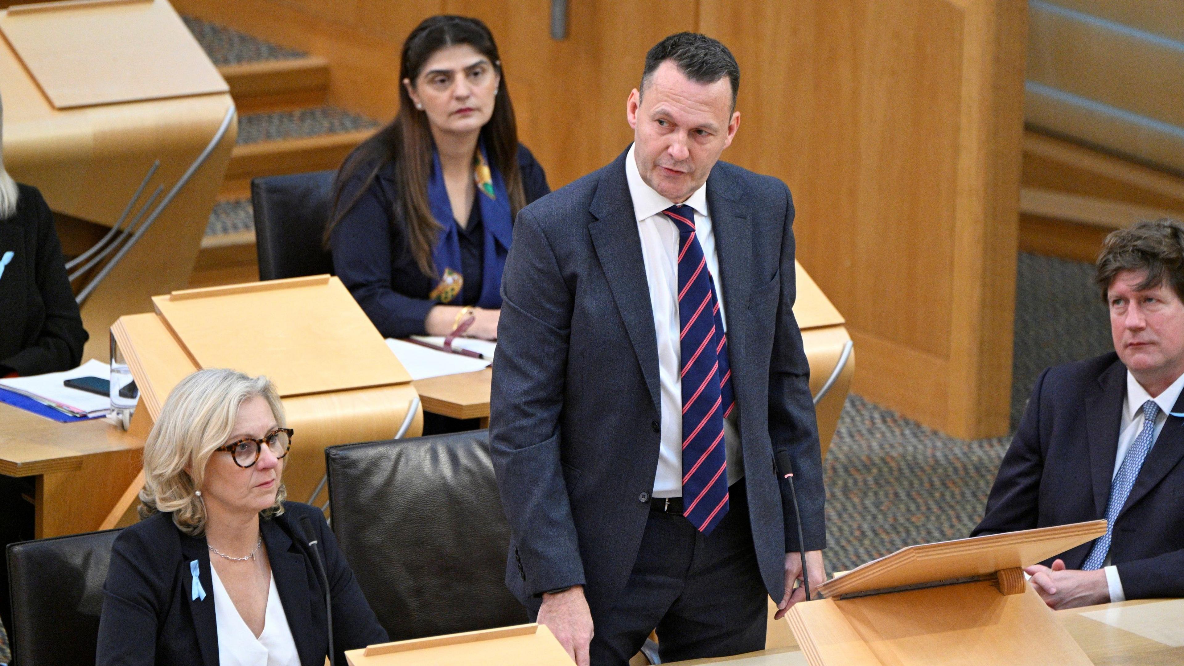 Russell Findlay standing up in parliament during FMQs. He is surrounded by his fellow MSPs