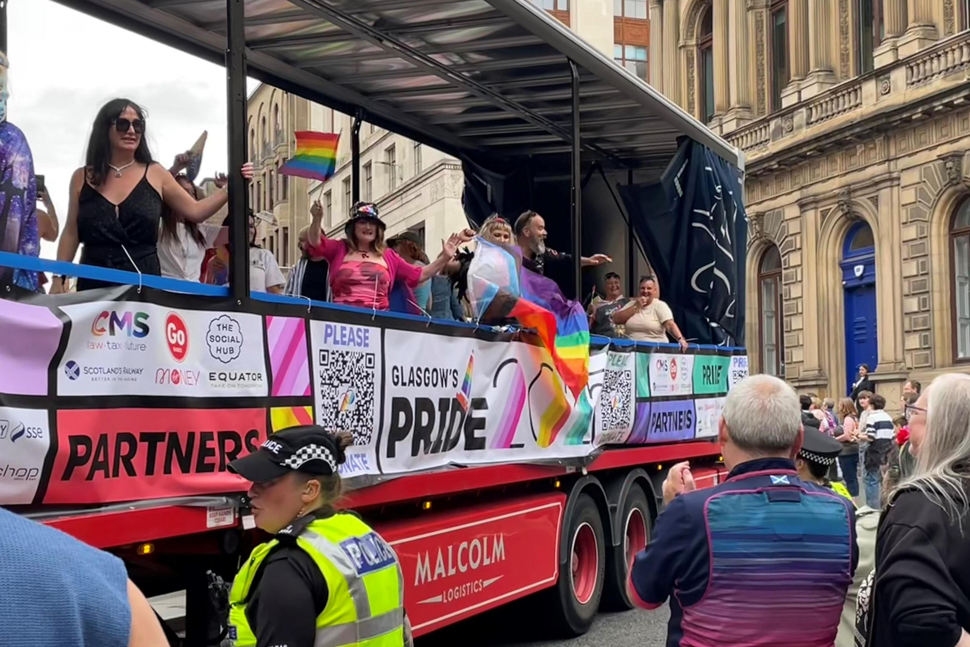 People in colourful clothes and flags on the back of an open-sided lorry that has Pride banners attached