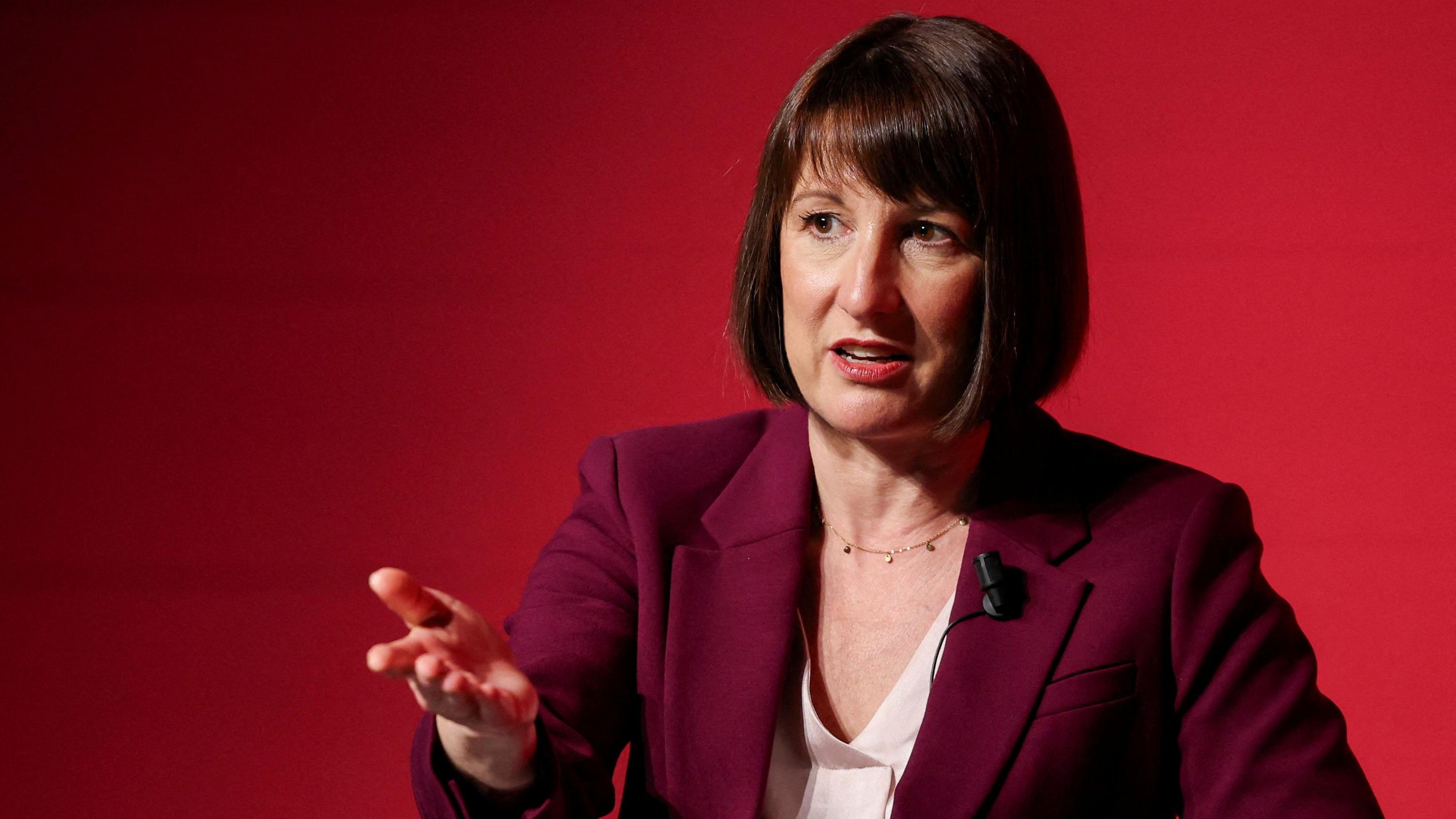 Rachel Reeves, sitting down for a TV interview, is wearing a plum-coloured suit jacket and a v-necked cream blouse underneath, with a microphone on her left lapel. She is sitting in front of a red background and gesturing with her right hand. 