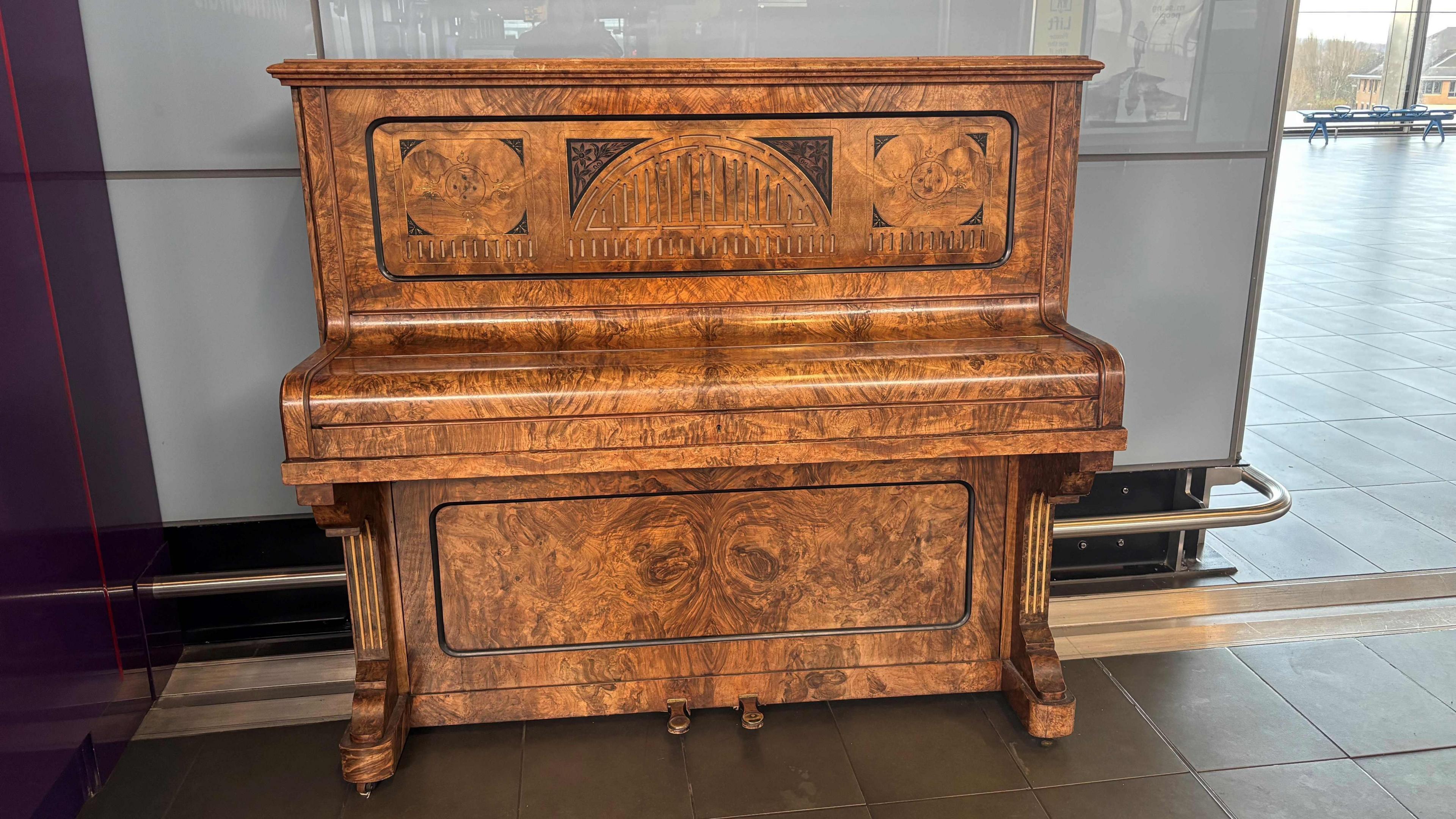 An upright piano made of light patterned wood, standing in a station for commuters to play.