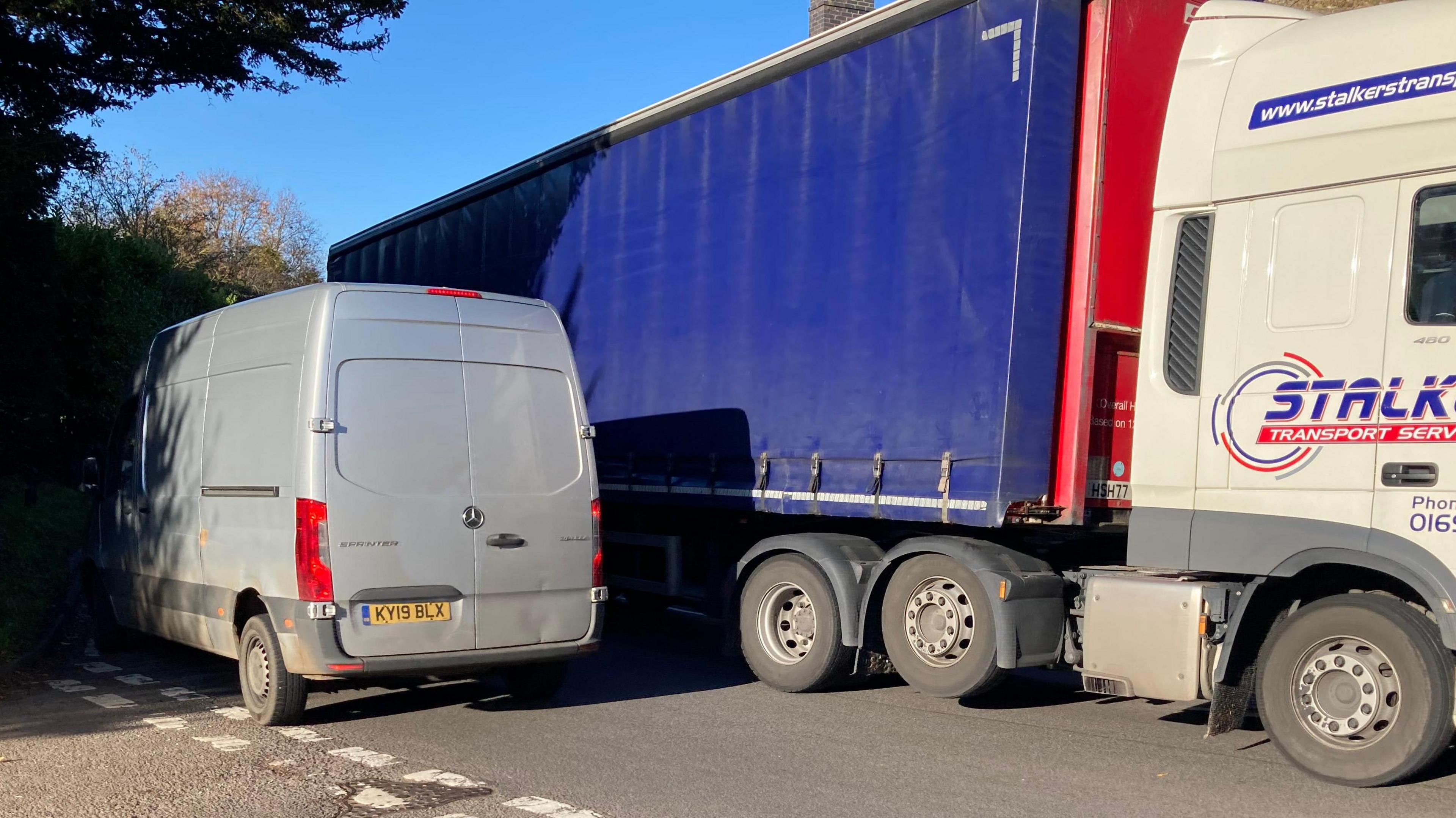 A grey van struggles to pass a long blue lorry on a bend in the road. 