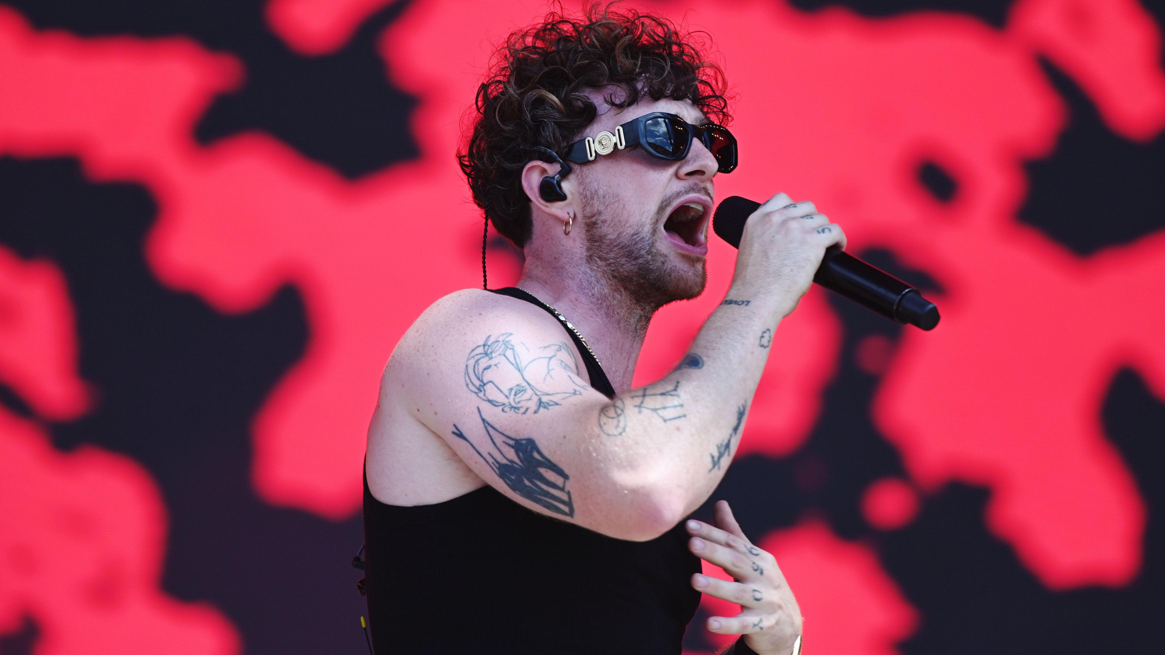 Tom Grennan singing into a microphone while performing live on stage at Glastonbury. He has short brown, curly hair and is wearing sunglasses, a blank tank top and has tattoos on his right arm and left knuckles.