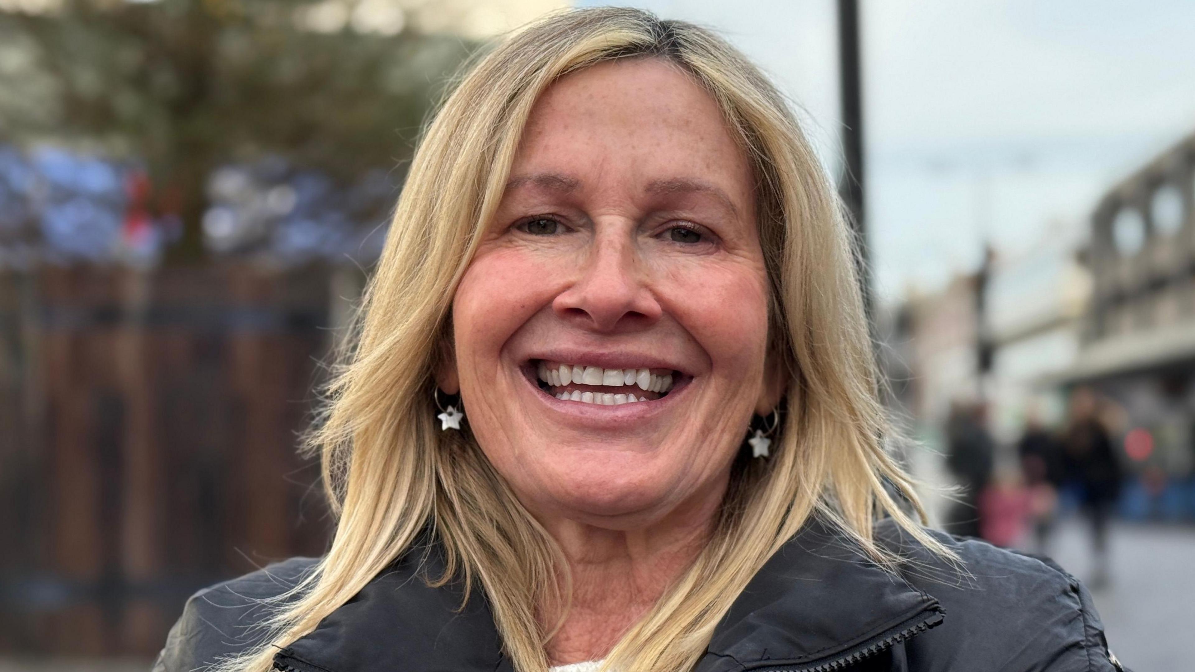 Marejka Darcy-Rampling is smiling at the camera while standing in front of the controversial tree. She has long blonde hair, star earrings and a black coat on. 