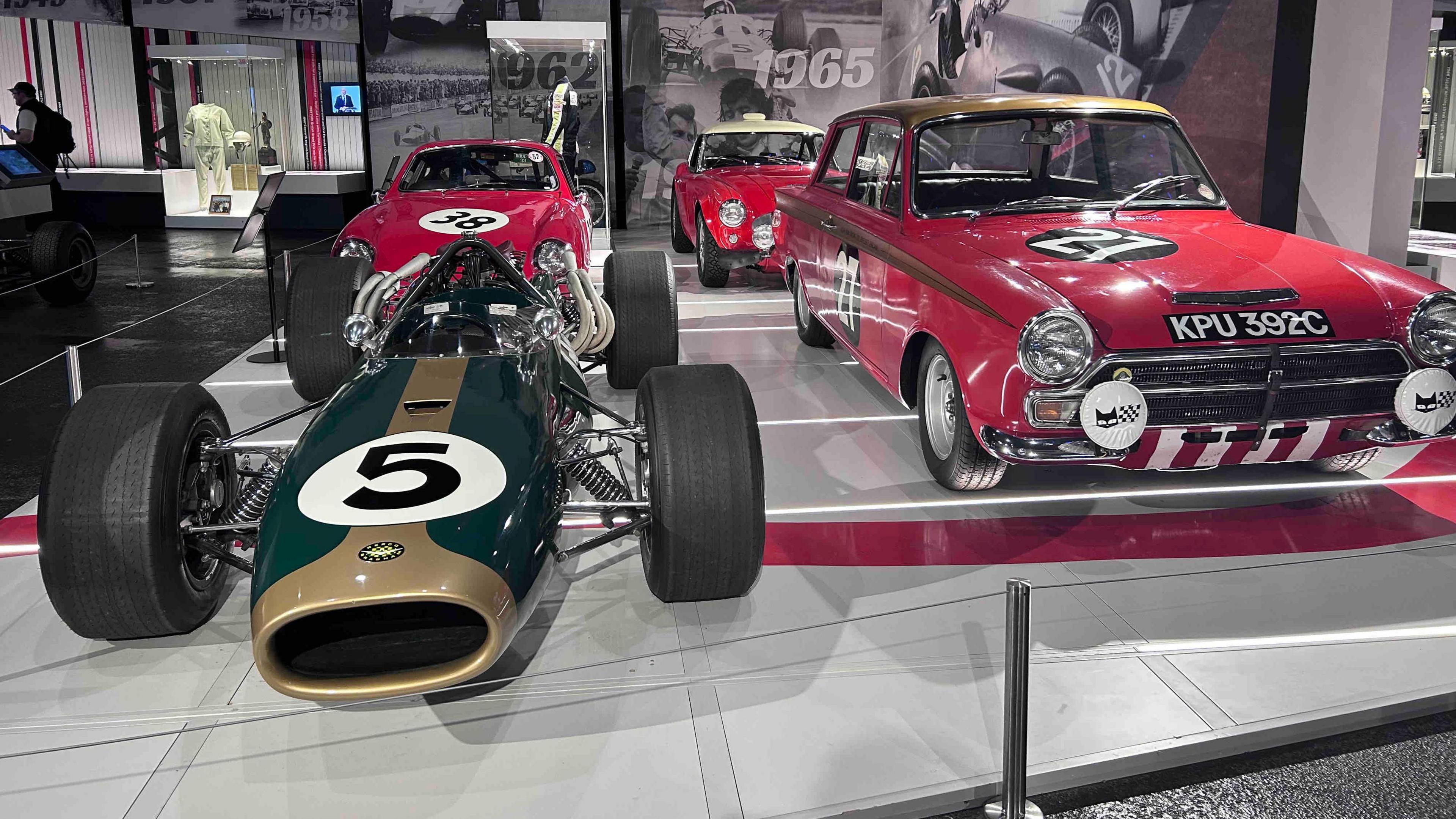 A dark green and gold racing car with a number 5 on the front. There is a red car behind it and two further red cars to the right. One has the number 27 in white writing on the bonnet. They are parked on a grey stage at an exhibition.