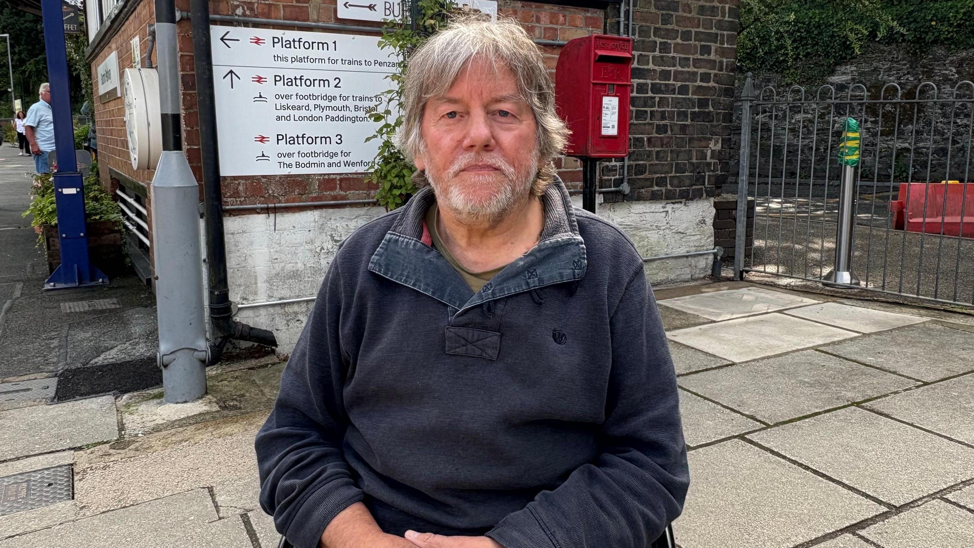 A man with long grey hair and a grey beard wearing a dark blue wide-collared top sitting in his wheelchair in front of a station building with a post box and sign behind him. 