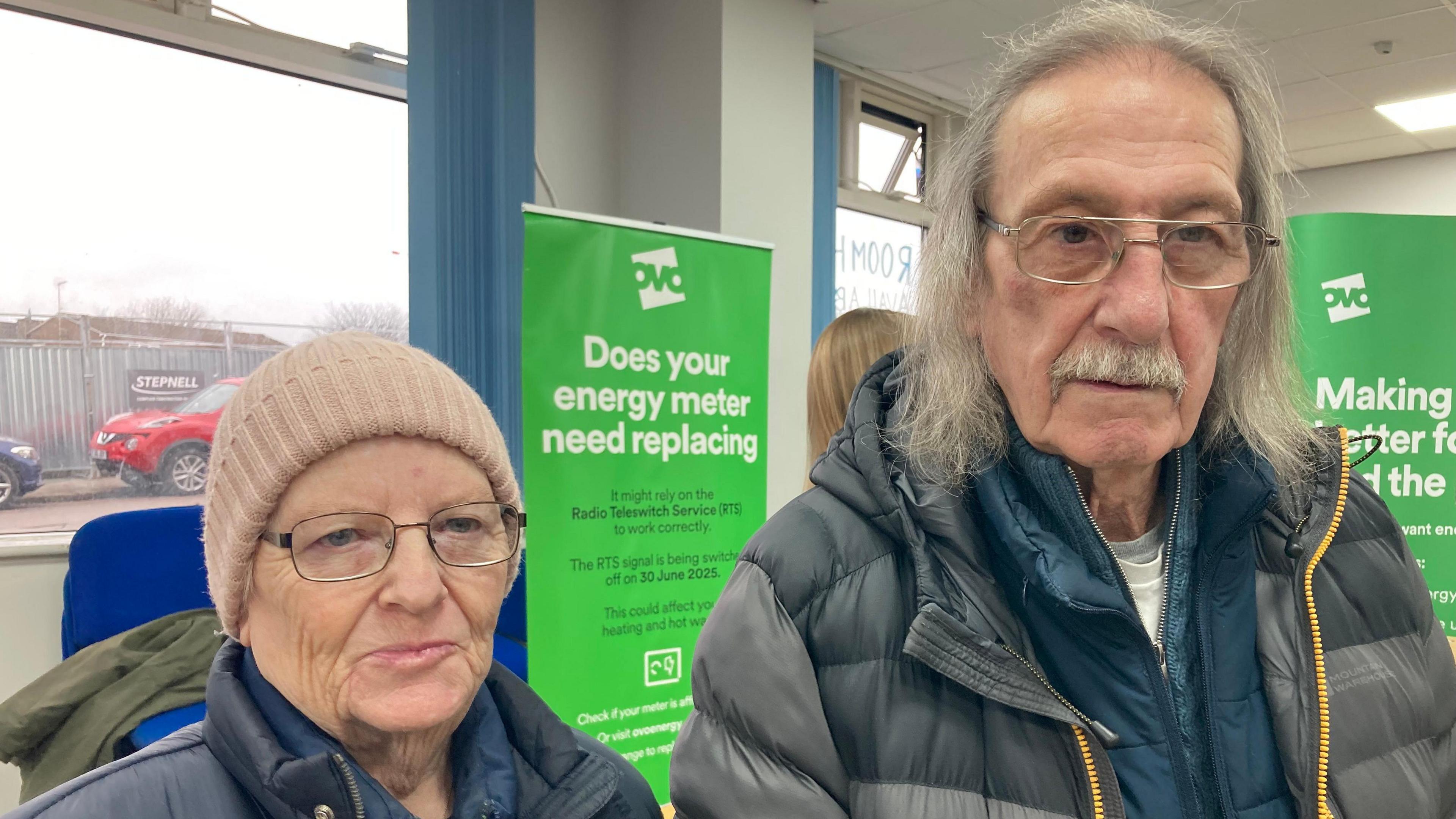 Glynis and Matthew Morgan standing side by side in front of a green Ovo energy banner informing visitors on energy meters. Glynis is wearing a blue padded coat, a light pink beanie and metal framed glasses. Matthew is wearing a blue jacket underneath a grey padded raincoat, and metal framed glasses. He has shoulder-length grey hair and a moustache. 