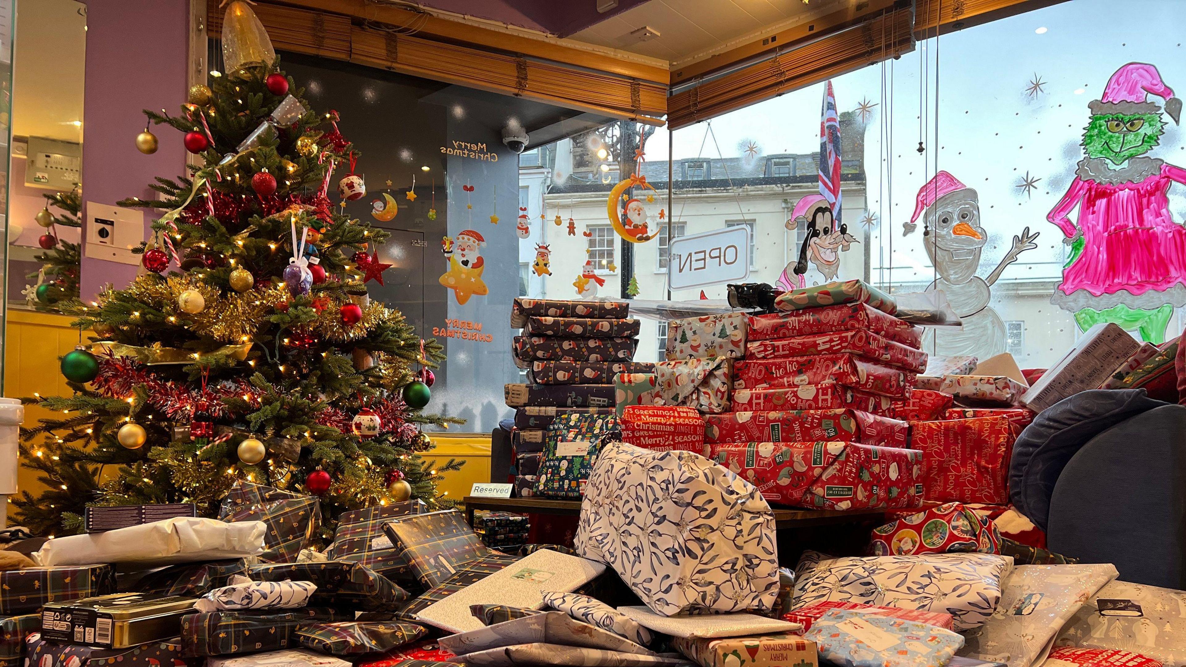 Presents are piled on top of each other in the cafe by its window and there's a Christmas tree to the left of the presents. On the windows are children's characters drawn on like Pluto, Olef and The Grinch.