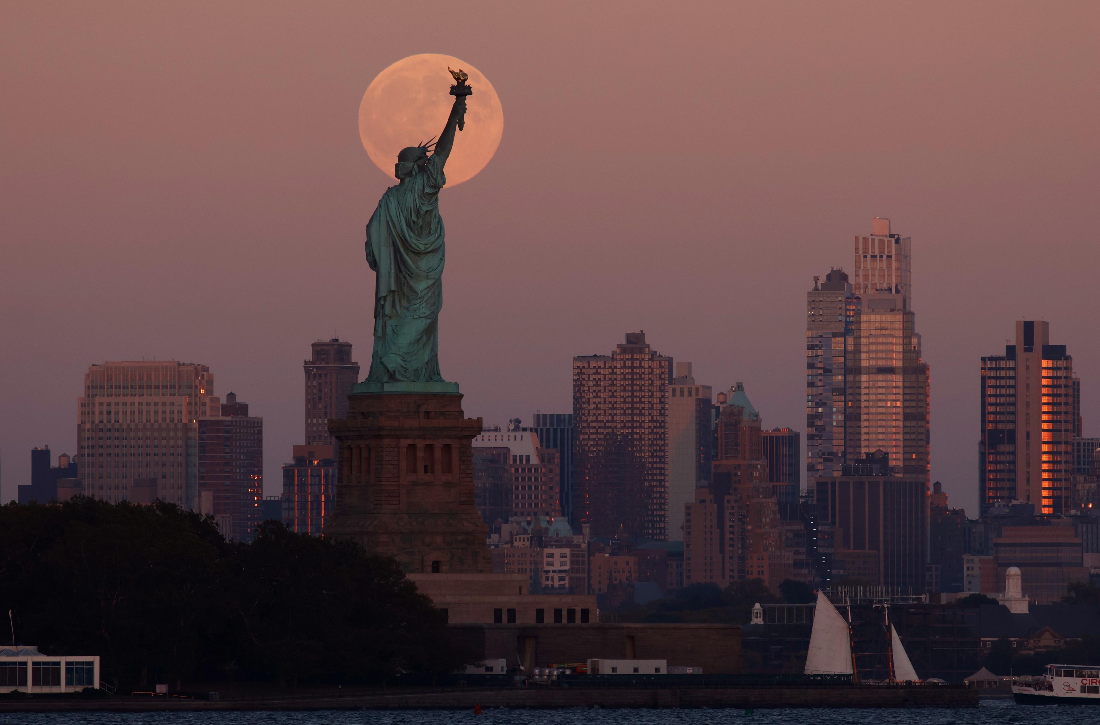 The supermoon appears behind the statue of liberty as the sun goes down over New York City, washing the city in a pinkish glow