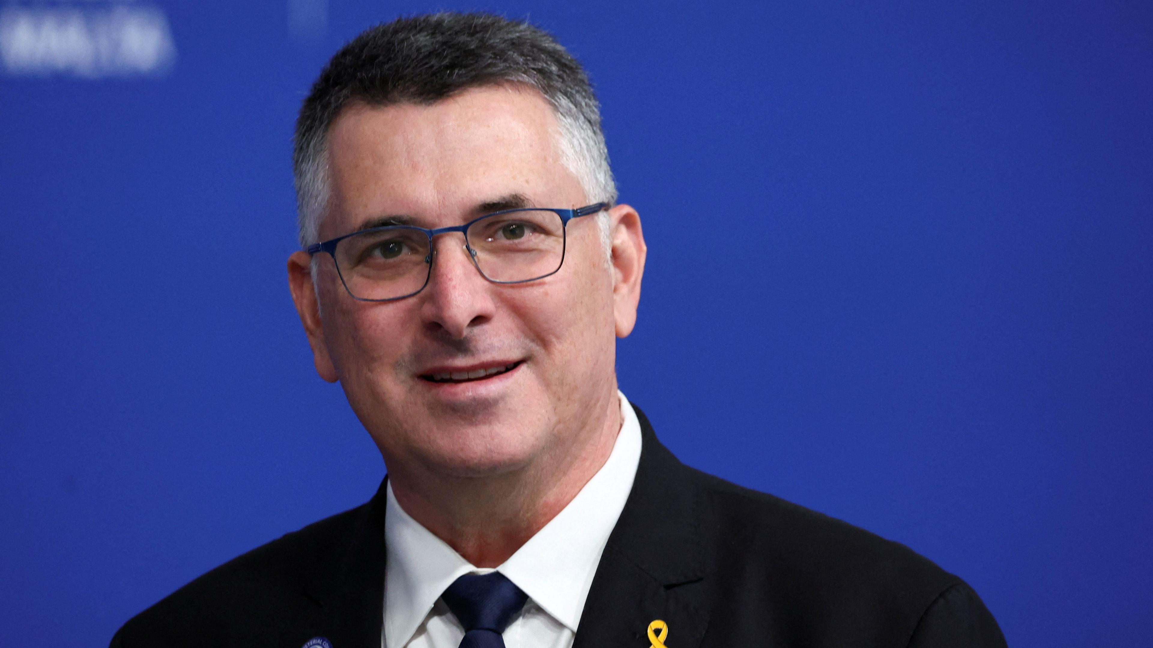 Israel's foreign minister, a male with grey hair wearing a dark coloured suit and tie with white shirt. Behind him is a blue background. 