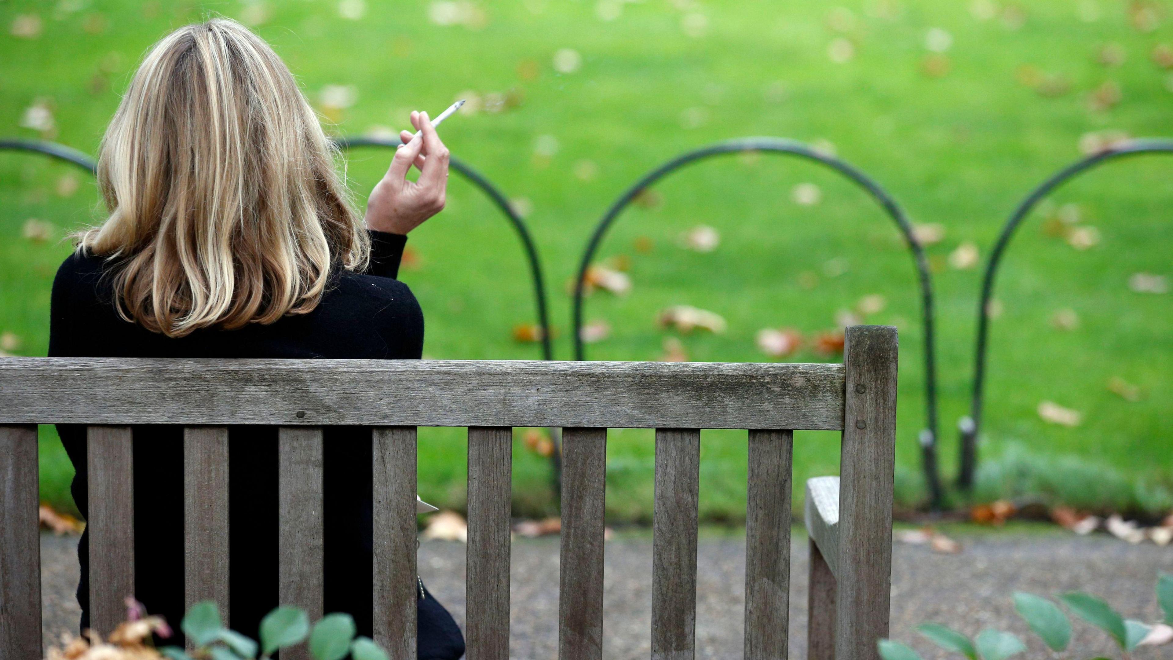 A woman with her back to the camera sitting on a park bench with a cigarette in her left hand. She has blonde hair just past her shoulders.
