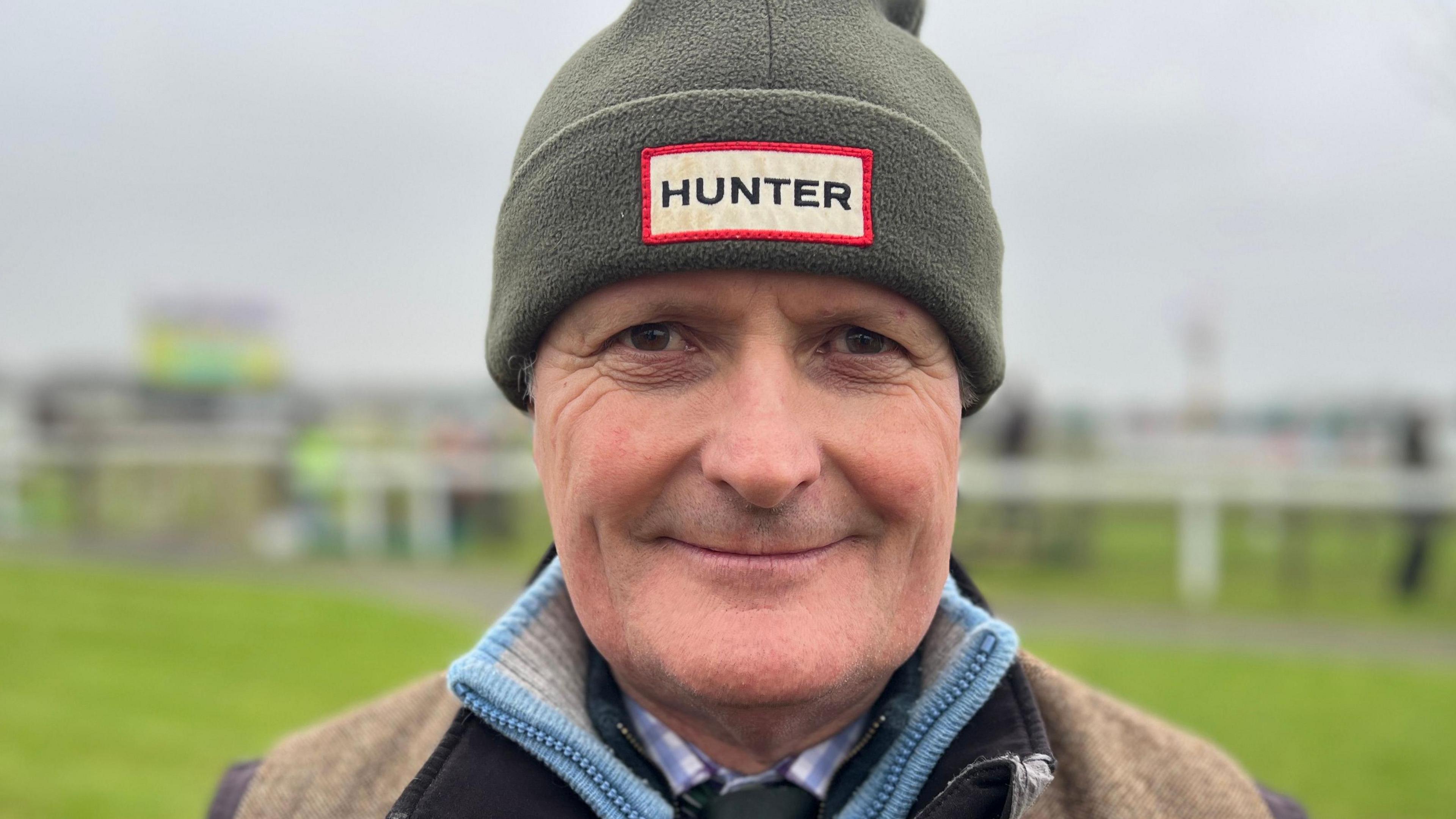 David Hunter is wearing a Hunter-branded green fleece hat, and a shirt and tie underneath two zipped jumpers and a country-style gillet. He is smiling and standing on the racecourse.