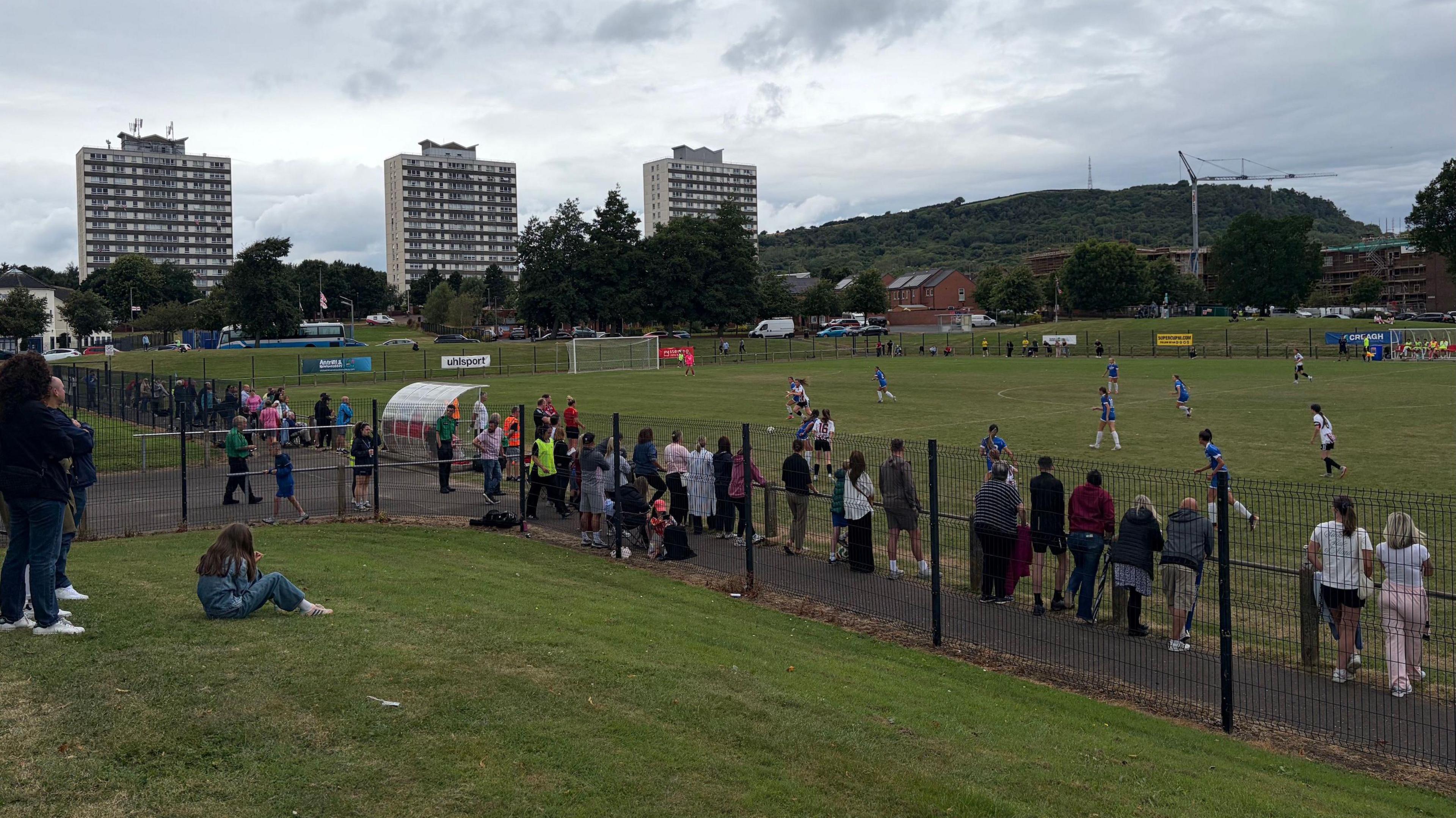 The Diamond in Rathcoole, a football pitch surrounded by a black fence. Parents watch the game between Crusaders in white and Surf Select in Blue. Three high-rise blocks of flats are to the left behind the pitch.