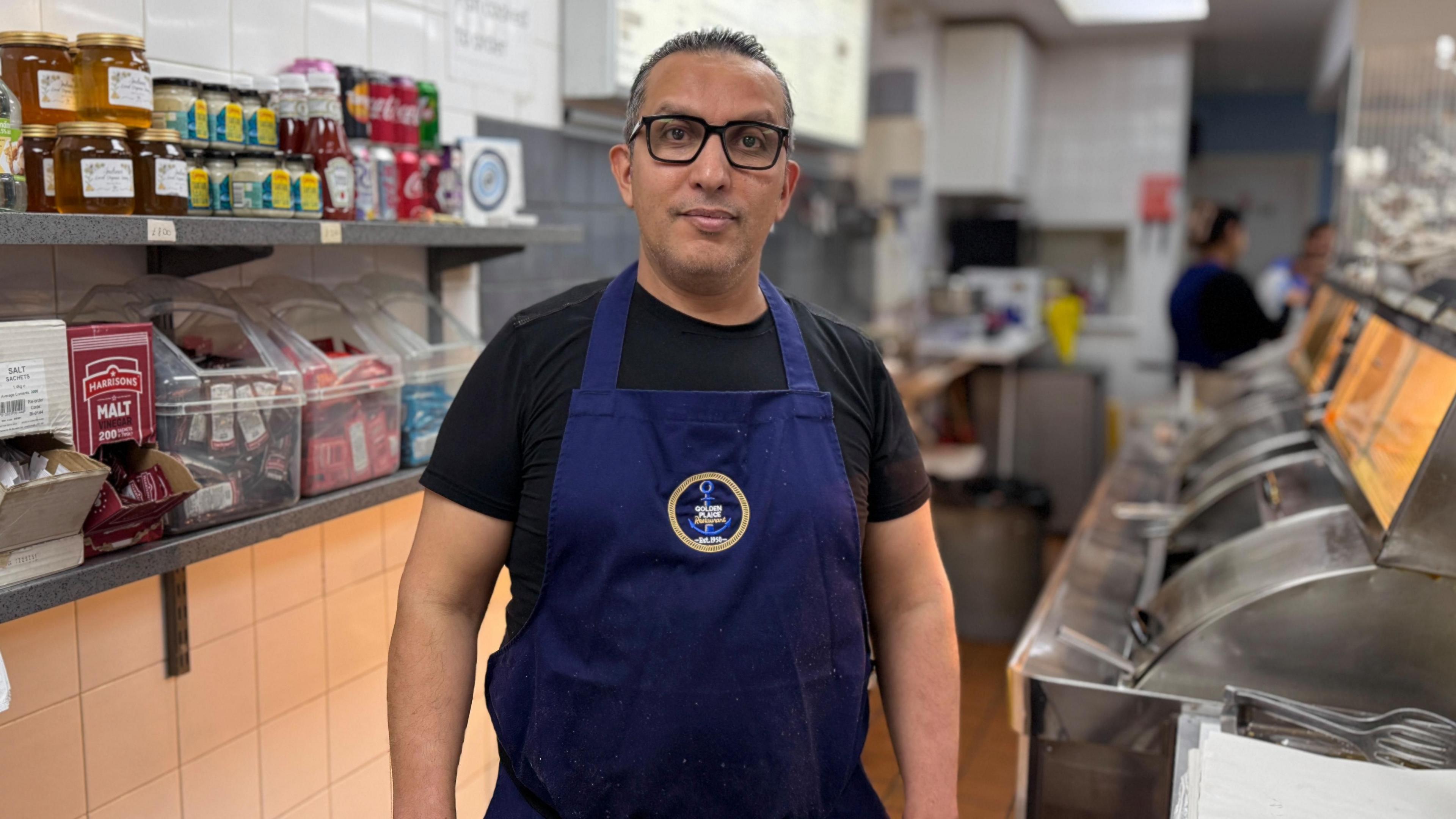 Aziz Chatt is stood behind the counter wearing a blue apron in his fish and chip shop