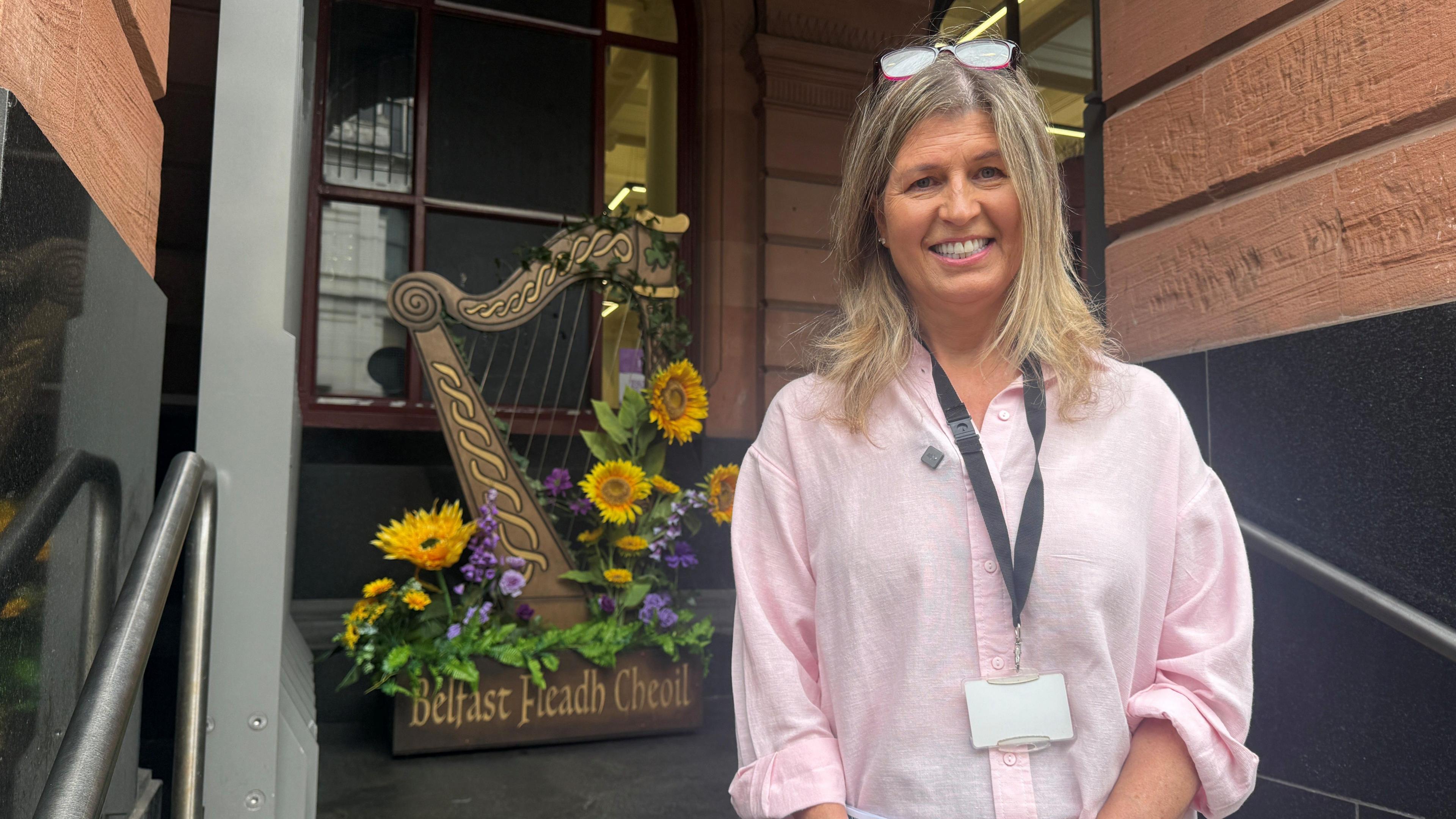 A woman with blonde hair smiling outside Central Library, a red brick building. She is wearing a pink blouse nad in the background is a large wooden harp sculpture with bright yellow flowers on it. It reads Belfast Fleadh Cheoil.