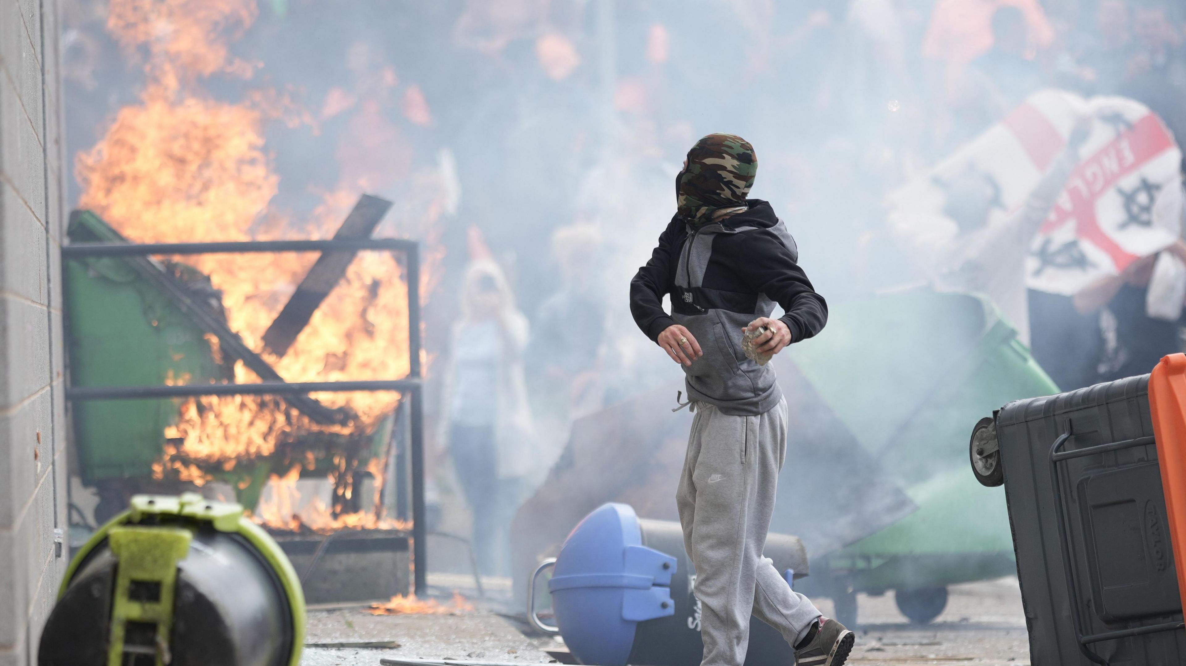 An image of rioting in Rotherham in August 2024. A protestor stands in the foreground of the image wearing grey tracksuit bottoms and a covering over his face and head. A crowd is in the background, behind a bin on fire next to a Holiday Inn Express