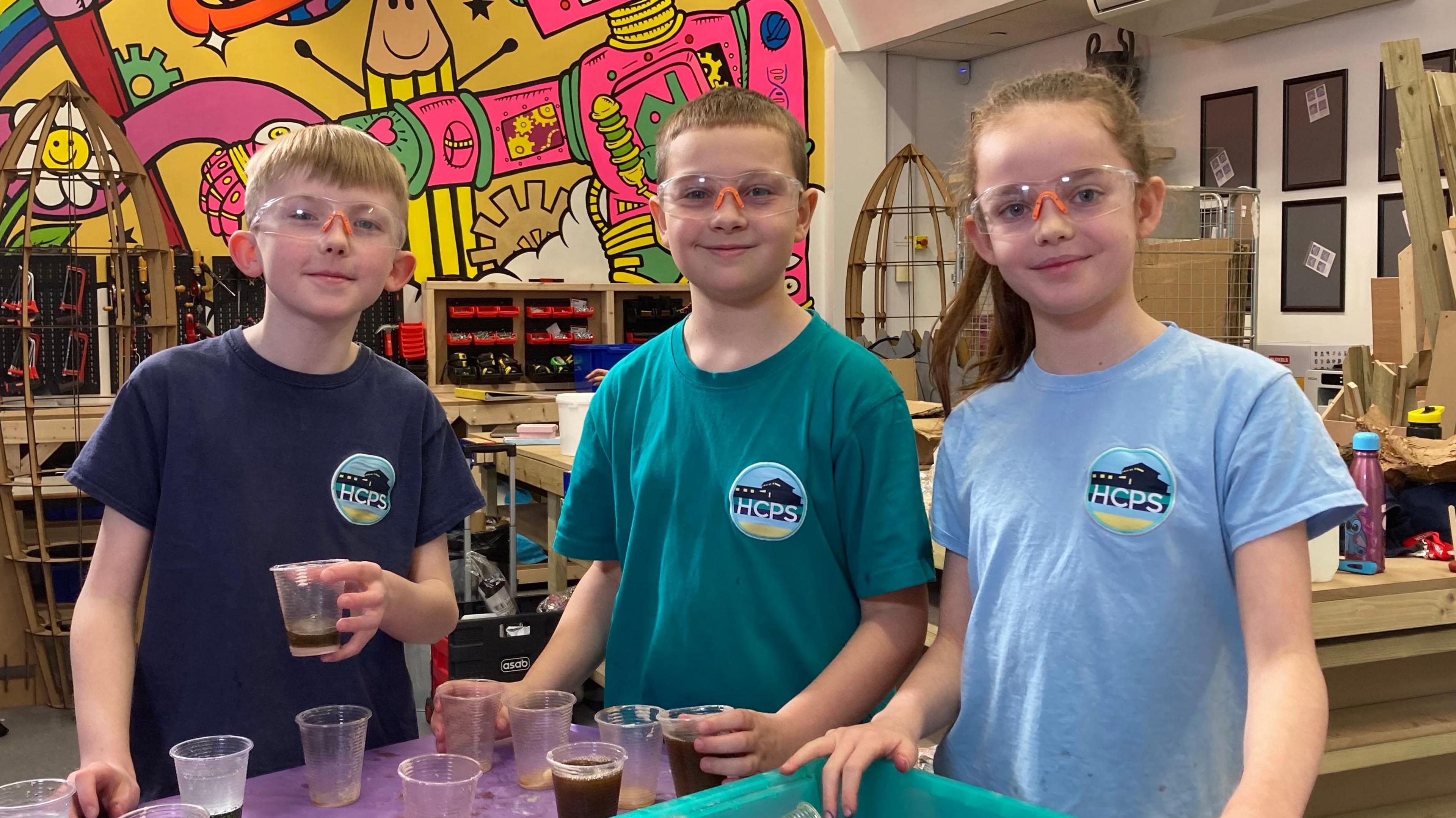 Three children stand in front of a table with a purple cover. The girl to the right holds on to a turquoise tray filled with plastic cups that have tipped over to the side. Greenish liquid has collected at the bottom of the tray. They all wear safety goggles.