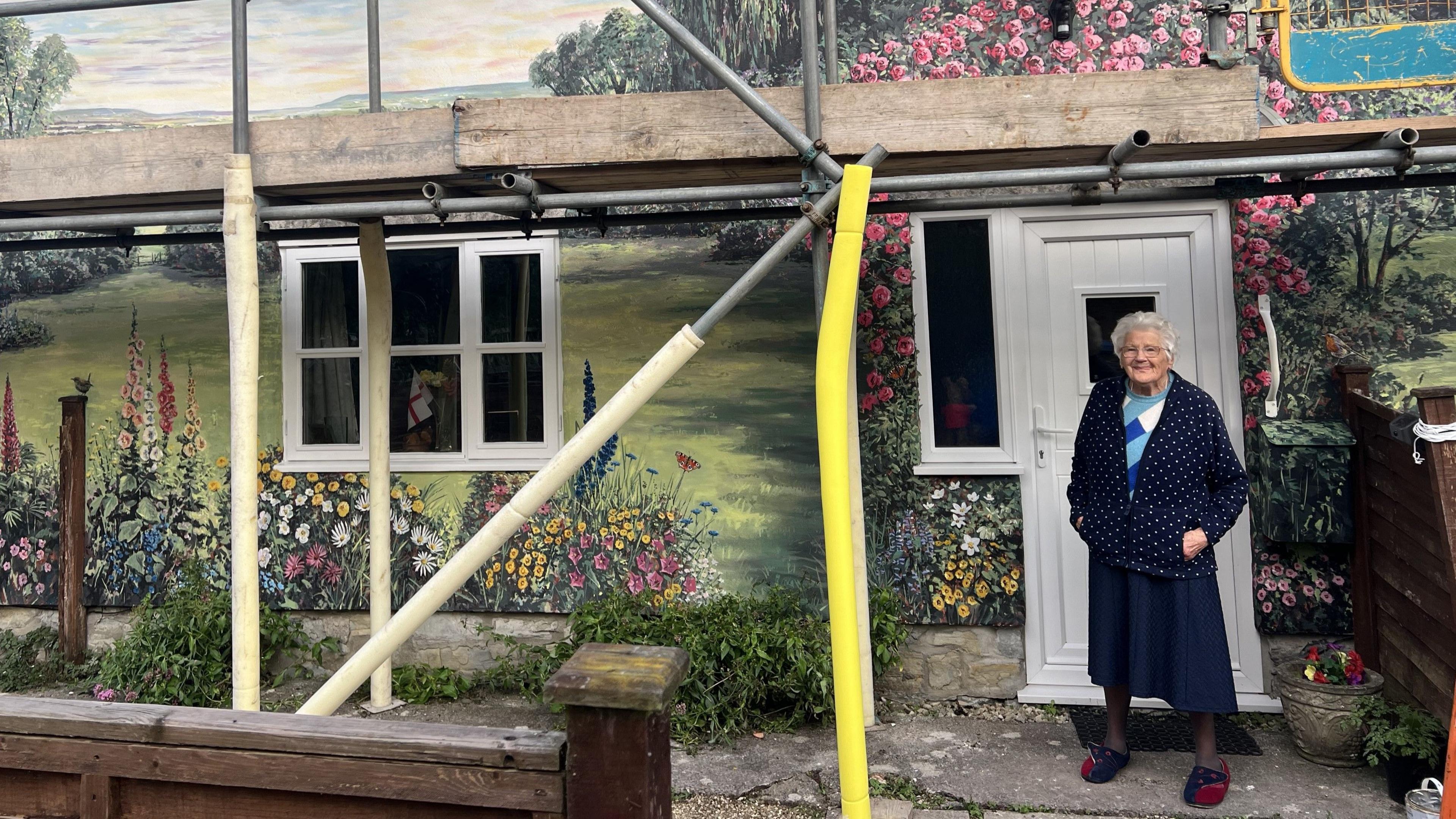 A woman stood in front of huge mural painted on a house. She is stood near to the house's white door. The wall is painted with grass, lots of colourful flowers, butterflies and there is a countryside landscape.