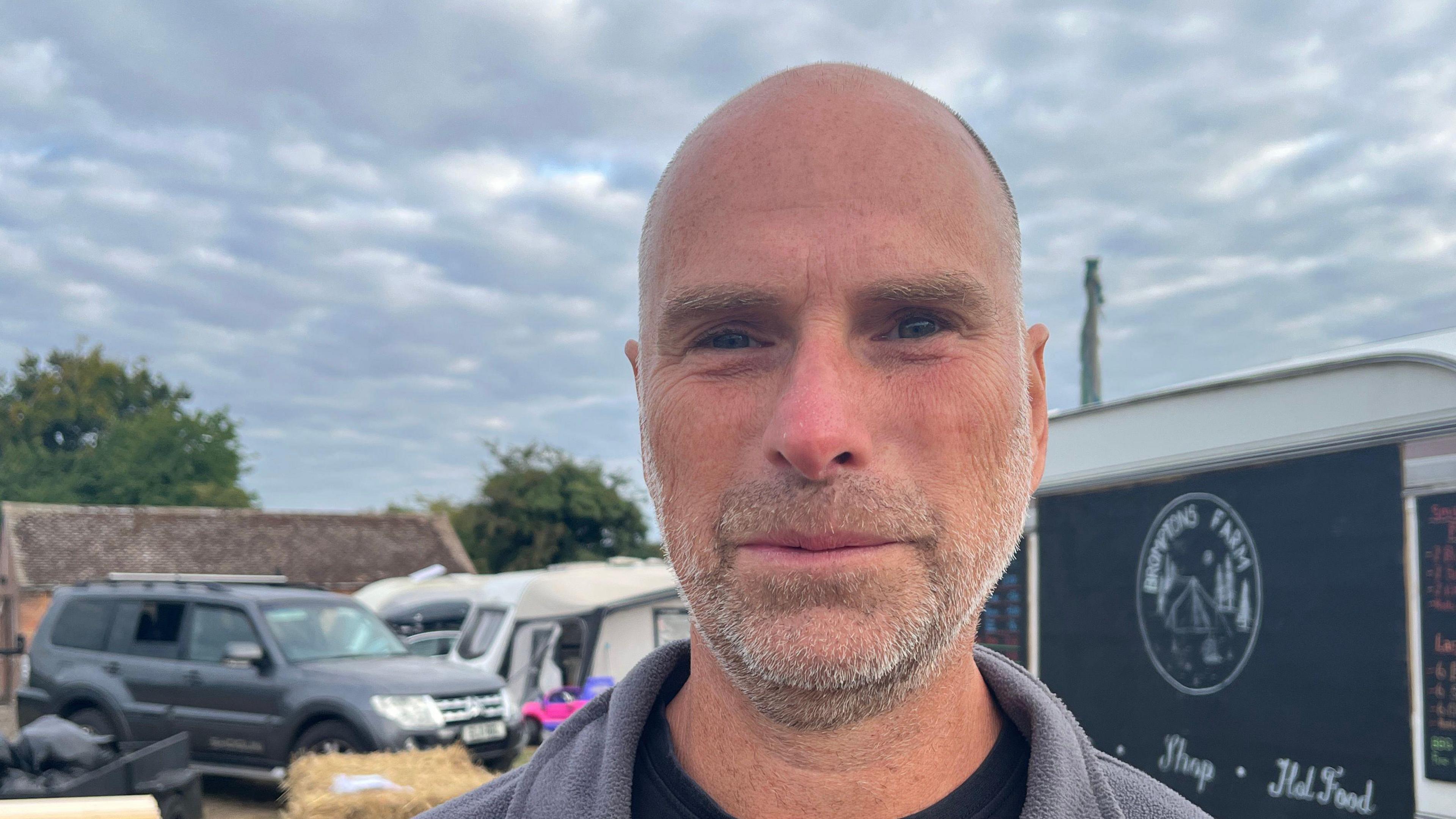 A close-up of Brenda Fisher. He is standing in front of a caravan, with other caravans and a 4x4 car in the background.