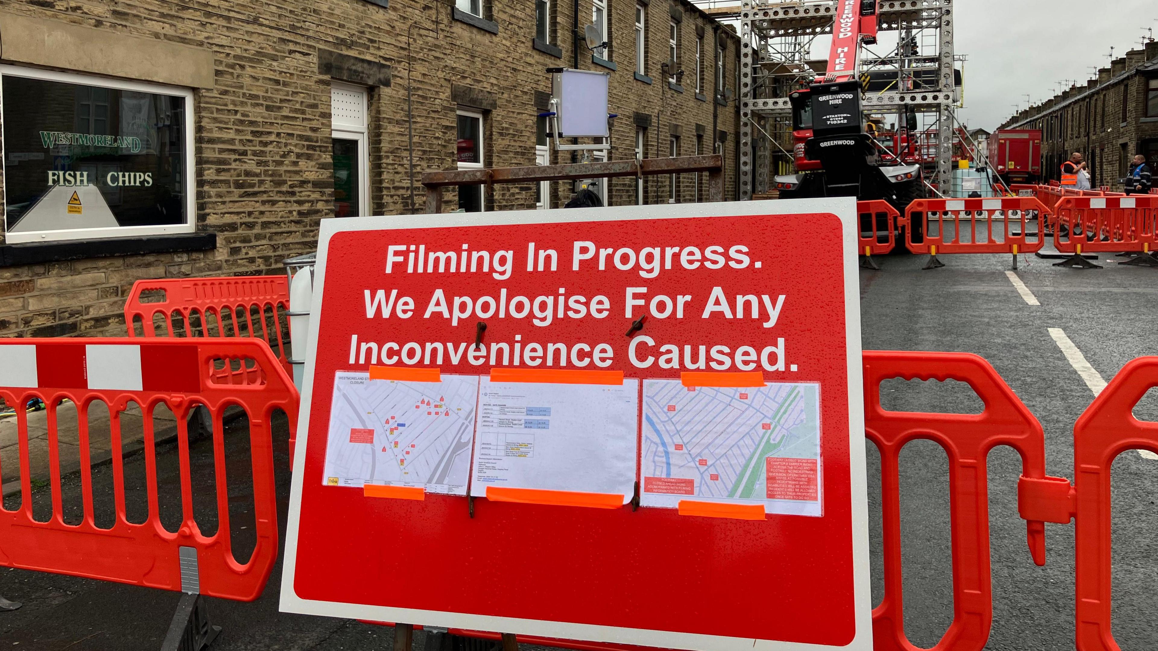 The image captures a street scene where filming is actively taking place. In the foreground, a sign reads: "Filming In Progress. We Apologise For Any Inconvenience Caused." Attached to the sign are three sheets, likely maps or diagrams, providing logistical details about the filming setup.
Behind the sign, a row of brick buildings lines the street, including a visible "Fish & Chips" shop. Orange safety barriers and construction equipment are positioned along the road.